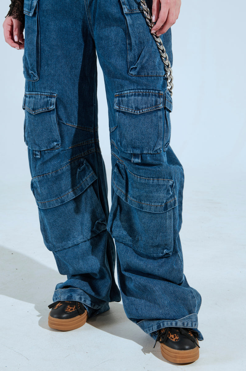 A person is rocking the "ALWAYS ON YOUR MIND DENIM JUMPSUIT IN BLUE," complete with wide legs, multiple pockets, and a chain accessory, perfectly paired with black and brown sneakers against a simple, light-colored background that enhances the casual vibe.