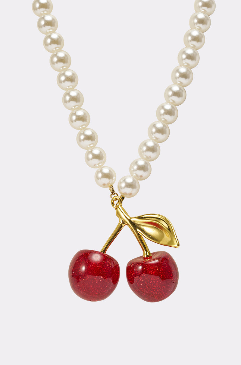 The POP MINE NECKLACE is a faux pearl necklace featuring a gold cherry pendant with two shiny red cherries and a gold leaf, displayed against a plain white background. 3