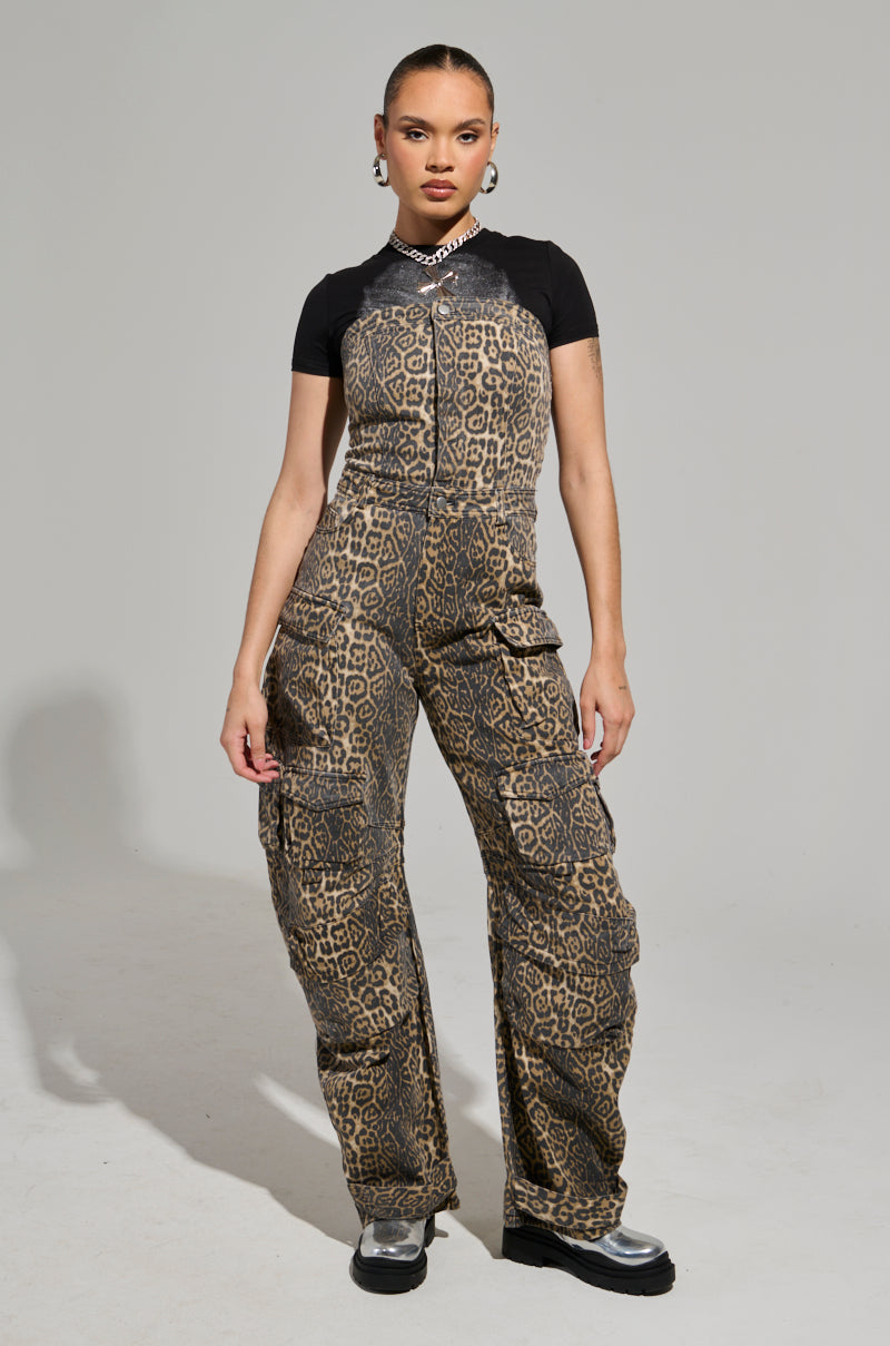 Dressed in the FIERCE ENERGY ONLY LEOPARD JUMPSUIT, a person channels intense energy with its wide leg silhouette while pairing it with a black T-shirt. They elevate the look with a chunky chain necklace and metallic silver boots against a plain gray backdrop that showcases their bold ensemble.
