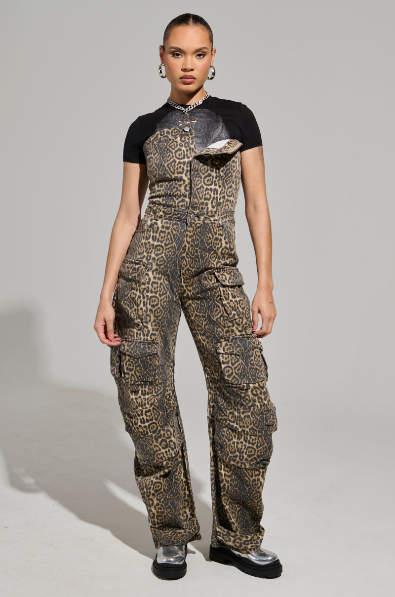 Exuding fierce energy, a person confidently flaunts the FIERCE ENERGY ONLY LEOPARD JUMPSUIT featuring a wide leg silhouette and black t-shirt sleeves. Hoop earrings frame their face, while metallic boots complete the look against a plain gray backdrop.