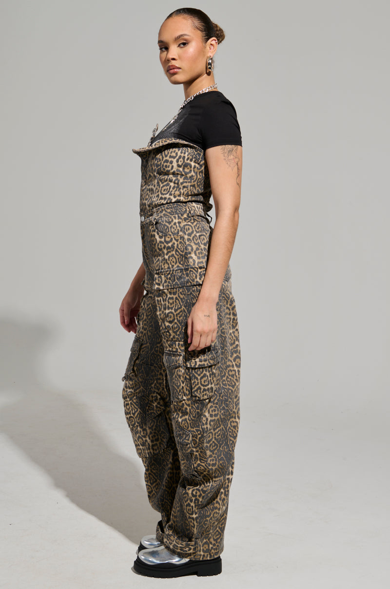 A person stands confidently in side profile, exuding fierce energy in a black top and the FIERCE ENERGY ONLY LEOPARD JUMPSUIT with a wide leg silhouette. Silver shoes glint beneath, while tattoos adorn their arms. The background is plain gray.
