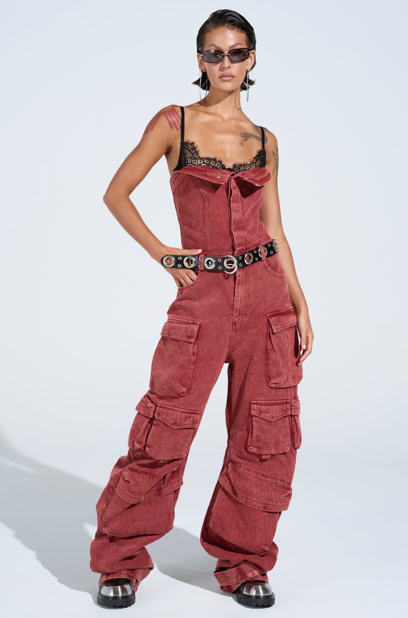 Donning dark sunglasses and short dark hair, a person exudes utility-chic style in the NOT INVITED CARGO JUMPSUIT, a red-wash denim creation featuring cargo pockets, black lace chest details, and a sleek black belt with silver rings against a plain background.