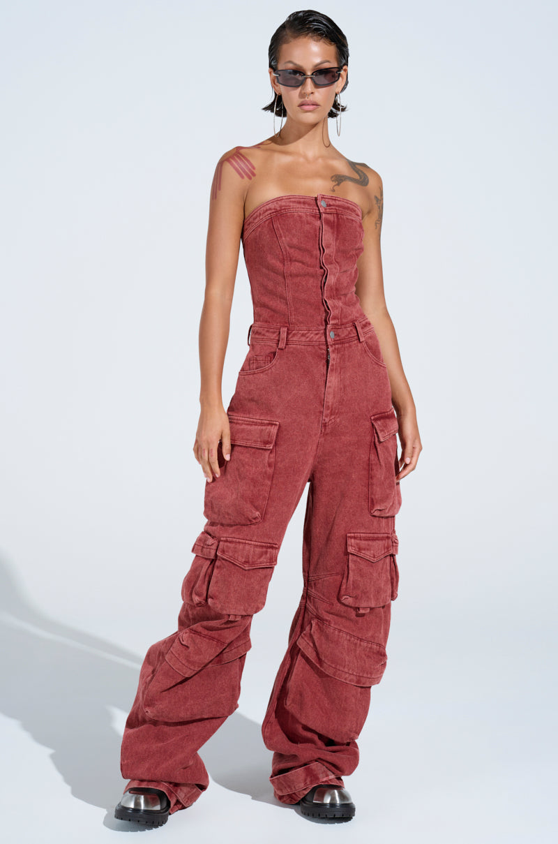 In a light setting, someone embodies utility-chic style in the NOT INVITED CARGO JUMPSUIT, a strapless red piece. Complemented by slicked-back hair, sunglasses, and black shoes with clear soles, they confidently represent modern fashion.