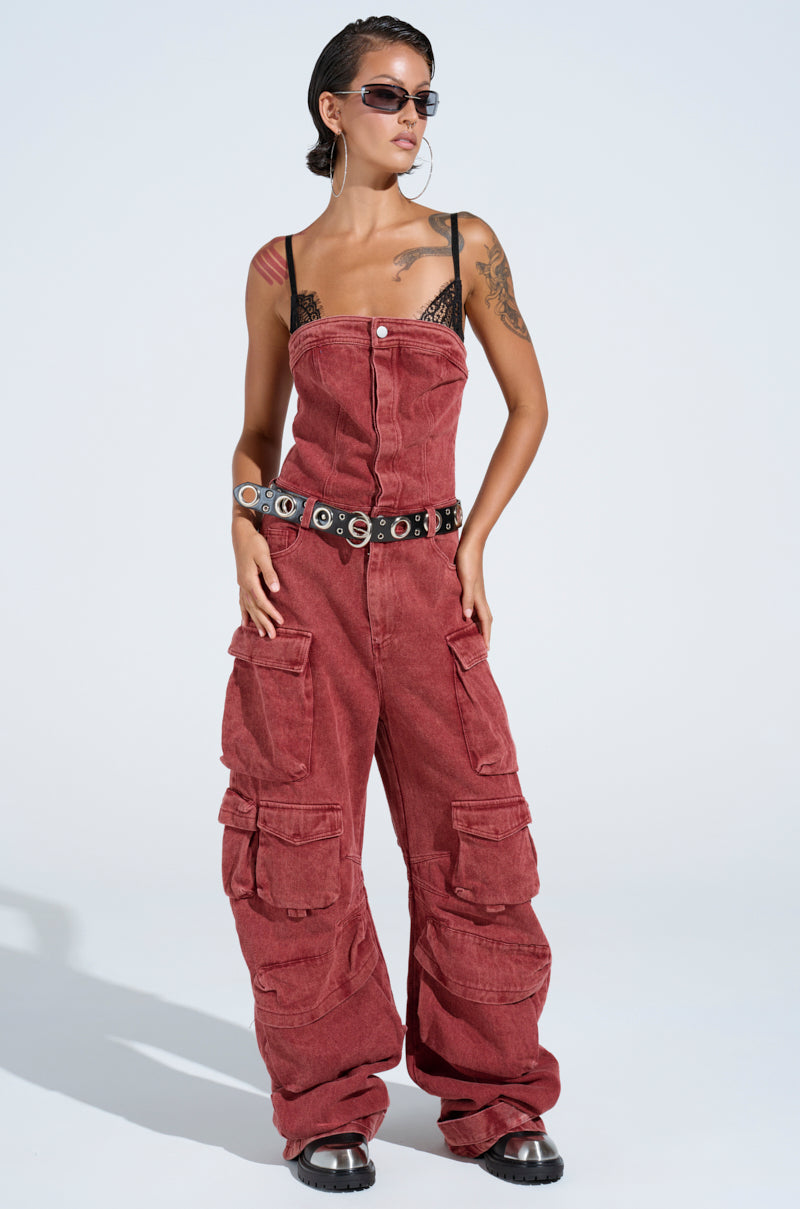 Clad in the NOT INVITED CARGO JUMPSUIT, this person exudes utility-chic flair. They pair it with a black lace top, sunglasses, a studded belt, and black shoes. Their hair is styled back to reveal a shoulder tattoo.