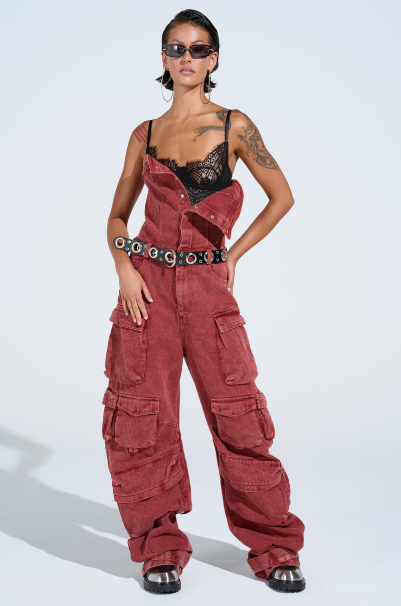 The NOT INVITED CARGO JUMPSUIT exudes utility-chic confidence with its oversized red-wash design, partially unzipped to reveal a black lace top. Paired with dark sunglasses, chunky black shoes, and a silver-studded belt, the look is completed with slicked-back hair showcasing tattoos.