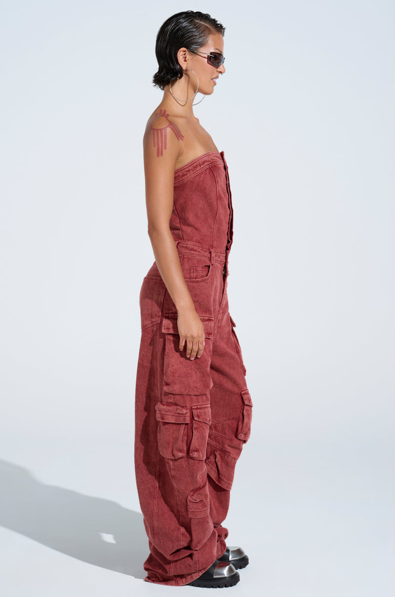 A person with short hair and hoop earrings wears the NOT INVITED CARGO JUMPSUIT, a strapless, oversized red-wash denim piece with multiple pockets. They sport sunglasses and exude utility-chic against a light gray background.
