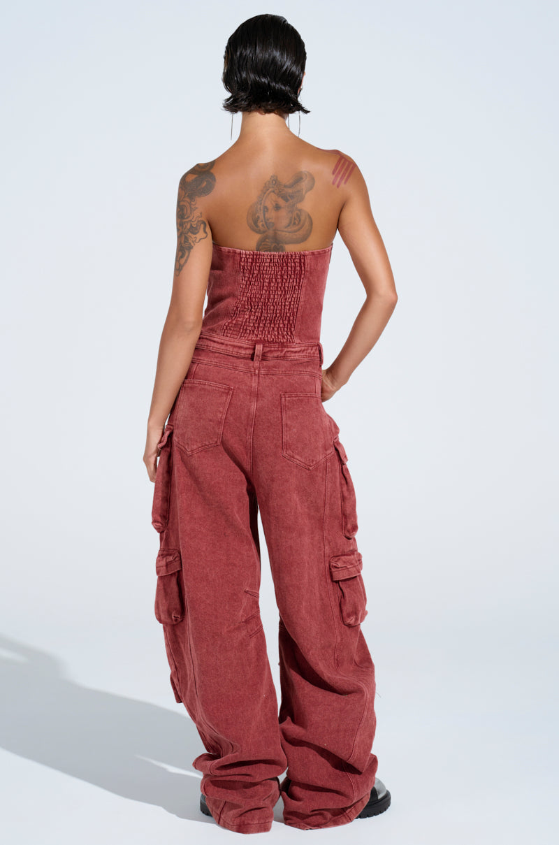 A person with short hair and tattoos has their back to the camera, wearing a NOT INVITED CARGO JUMPSUIT in dusty red for a utility-chic vibe, complemented by black shoes.