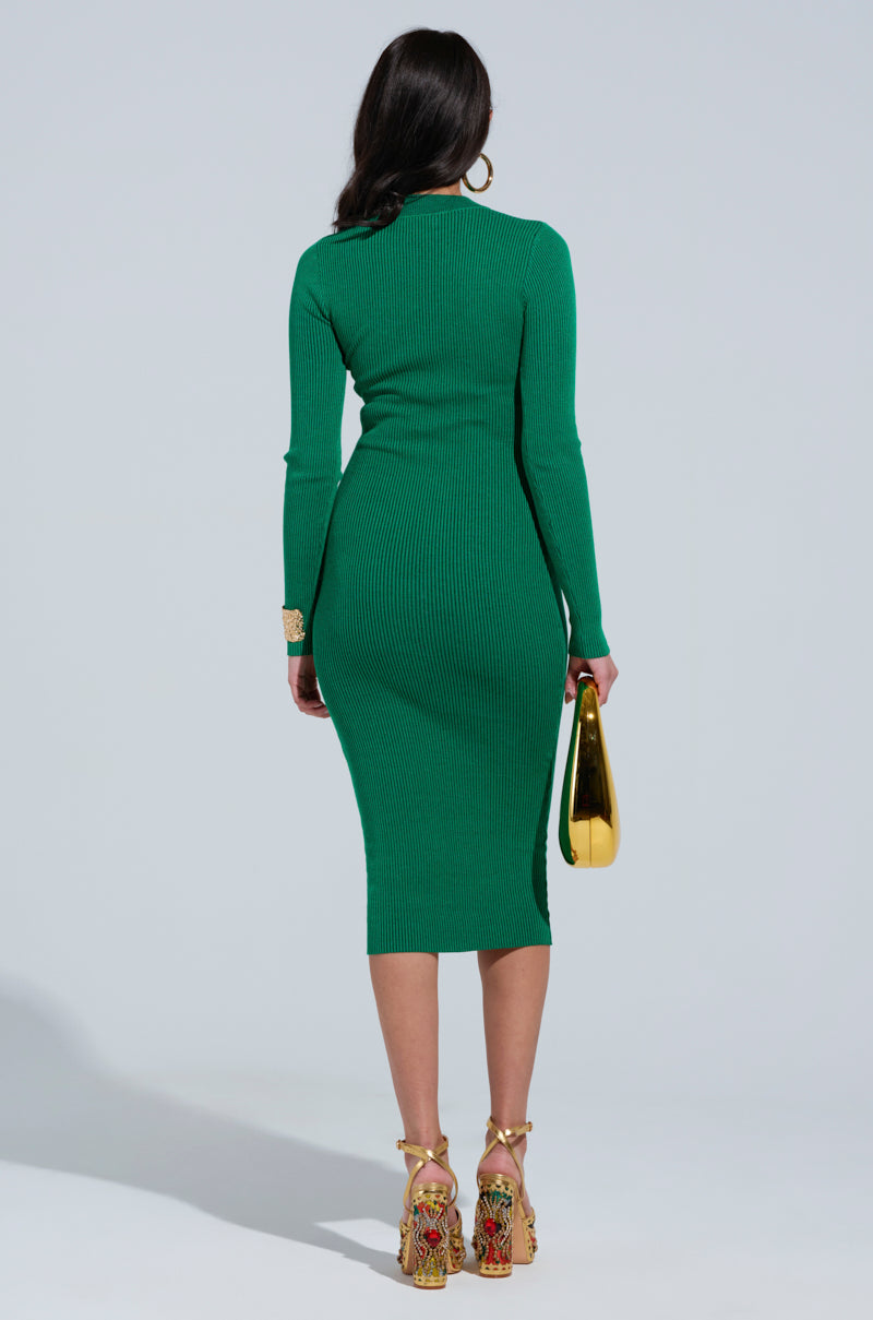 A woman stands facing away, wearing the ABOUT YOU MIDI DRESS IN GREEN and ornate high-heeled sandals. The bodycon fit accentuates her silhouette as she holds a gold, pear-shaped clutch in her left hand. Her long dark hair cascades down her back against the plain light gray background.