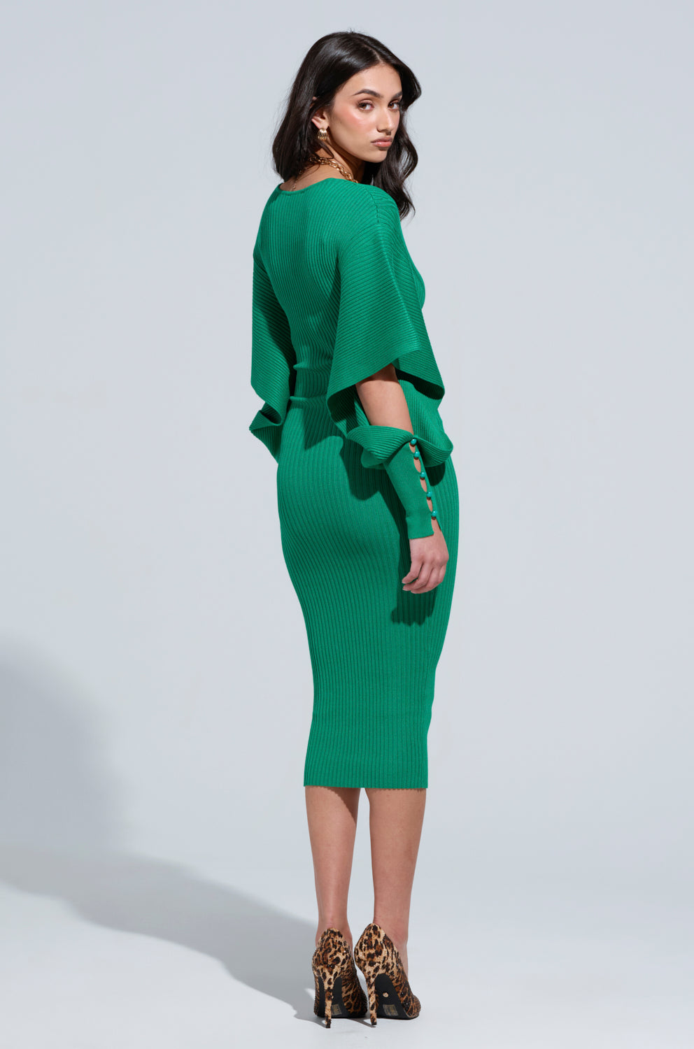 A woman stands in profile wearing the ONE OF YOU SWEATER MIDI DRESS, a ribbed knit green dress featuring ruffled sleeves. She pairs it with leopard print high heels and looks over her shoulder against a plain light gray backdrop.