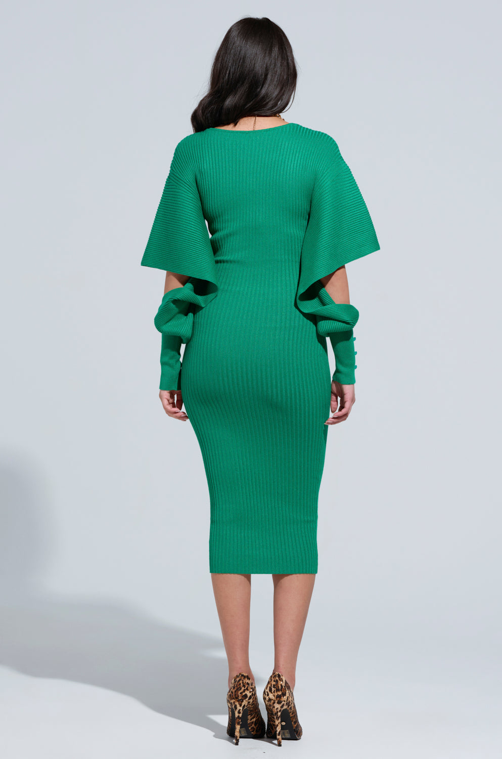A person with long black hair is wearing the ONE OF YOU SWEATER MIDI DRESS in green, featuring wide sleeves and matching arm warmers. They are standing with their back to the camera, highlighting leopard print high heels against a light gray background.