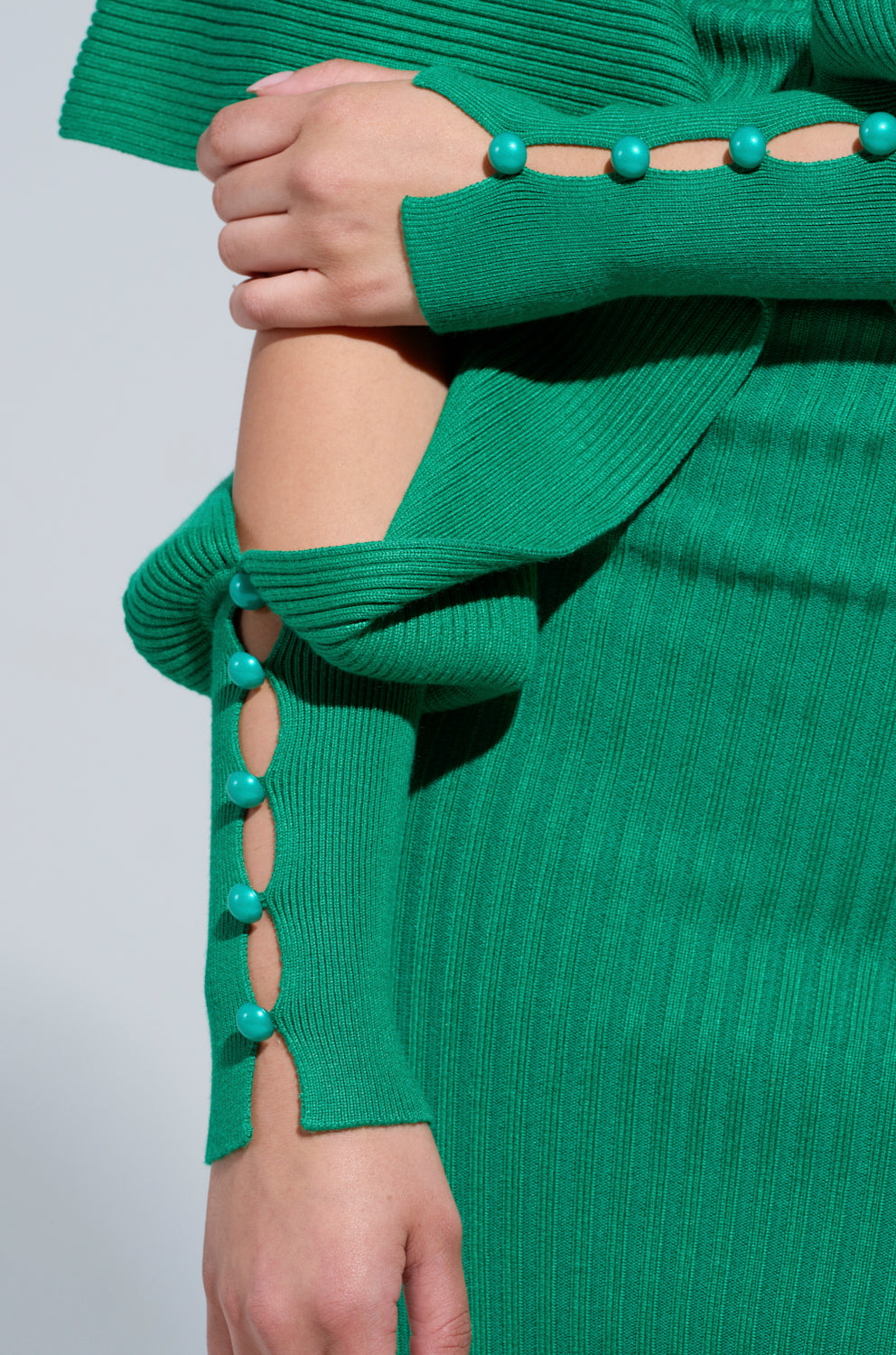 The individual is dressed in the ONE OF YOU SWEATER MIDI DRESS, a green ribbed knit sweater and skirt set adorned with large buttons along the sleeves and skirt, featuring a chic layered design at the cuffs. They hold their arm with one hand, elegantly highlighting the dress's sophisticated style.