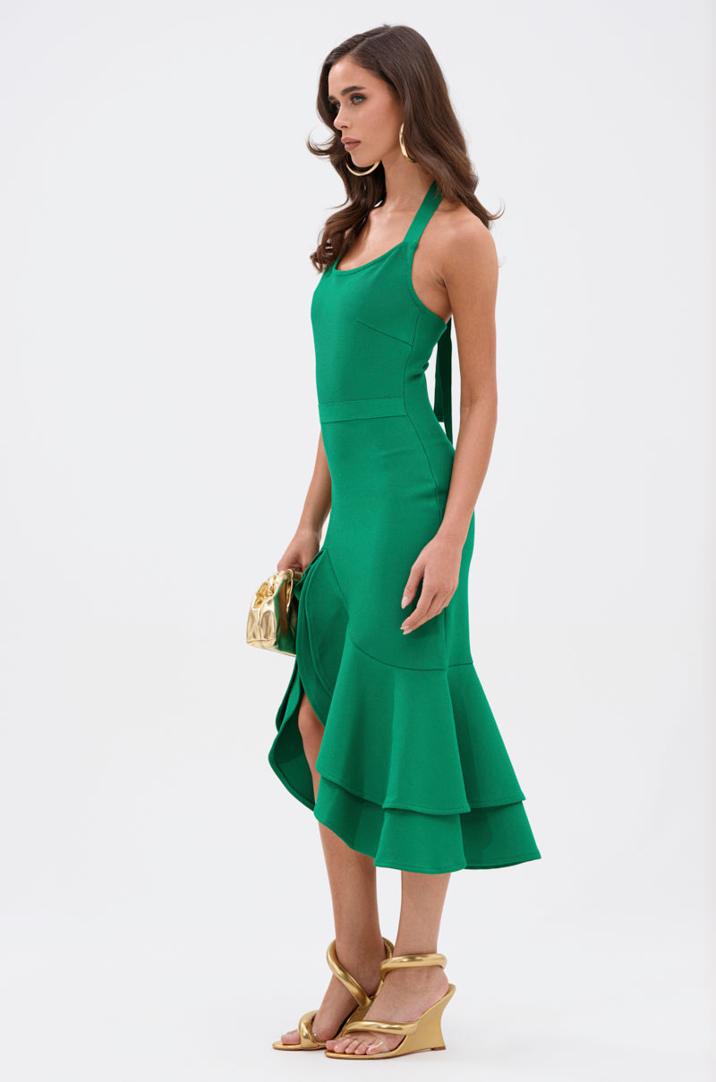 Wearing the LUCY MIDI BANDAGE DRESS IN KELLY GREEN, a woman holds a gold clutch, pairing strappy beige wedge sandals with loose waves as she poses elegantly against a plain white background.