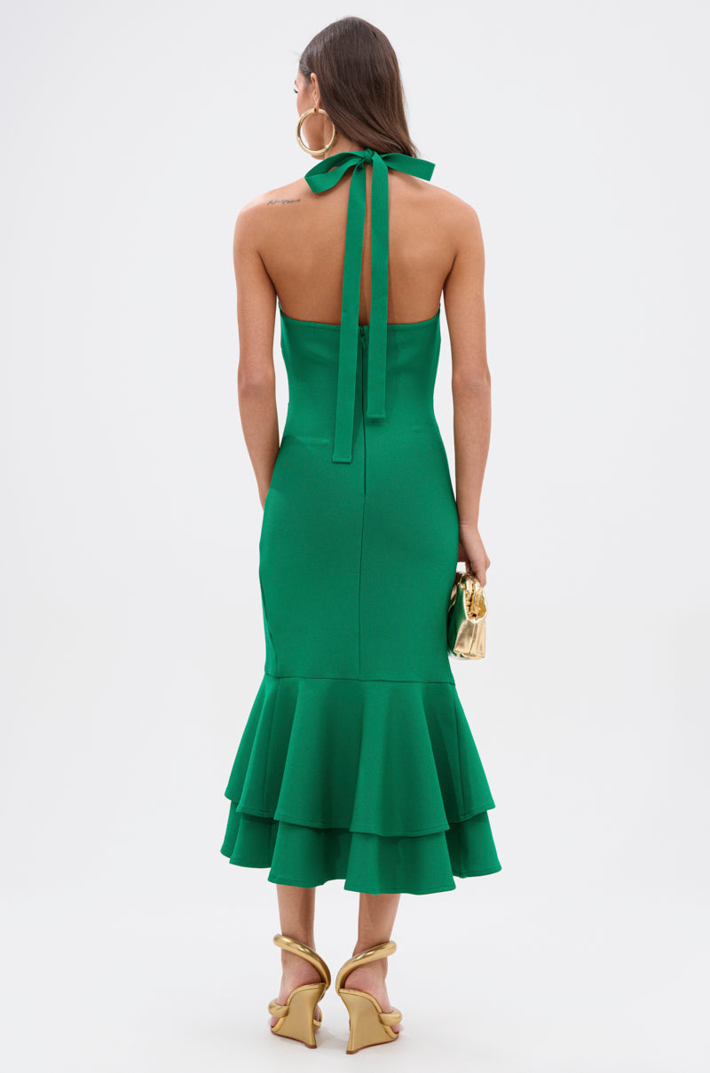 A woman, seen from behind, wears the LUCY MIDI BANDAGE DRESS IN KELLY GREEN featuring a halter neck, backless style, and ruffle accents. She accessorizes with a gold clutch, gold heels, and large hoop earrings for a chic bodycon look.