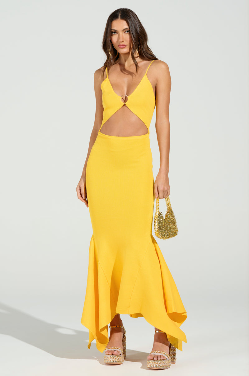 A woman holds a small woven bag and wears the "LOOKING FOR FUN KNIT MAXI DRESS IN YELLOW," featuring a cutout and flared asymmetrical hem. She pairs it with platform sandals against a plain, light-colored backdrop.