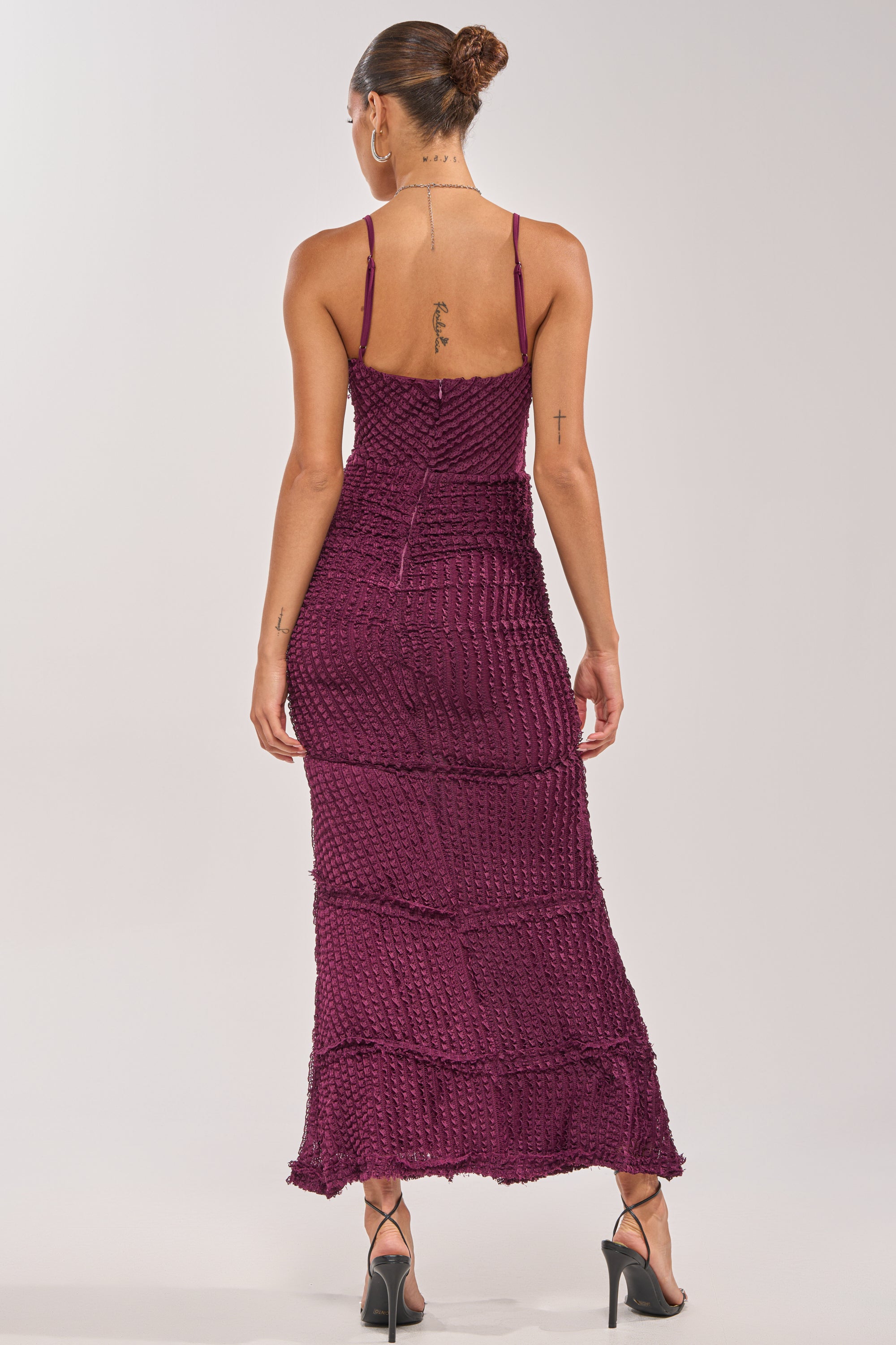A woman with her hair in a bun and glasses stands facing away, wearing the PLUS ONE MAXI DRESS IN PLUM with thin straps, paired with black high-heeled sandals. Tattoos are visible on her back and arm. 3