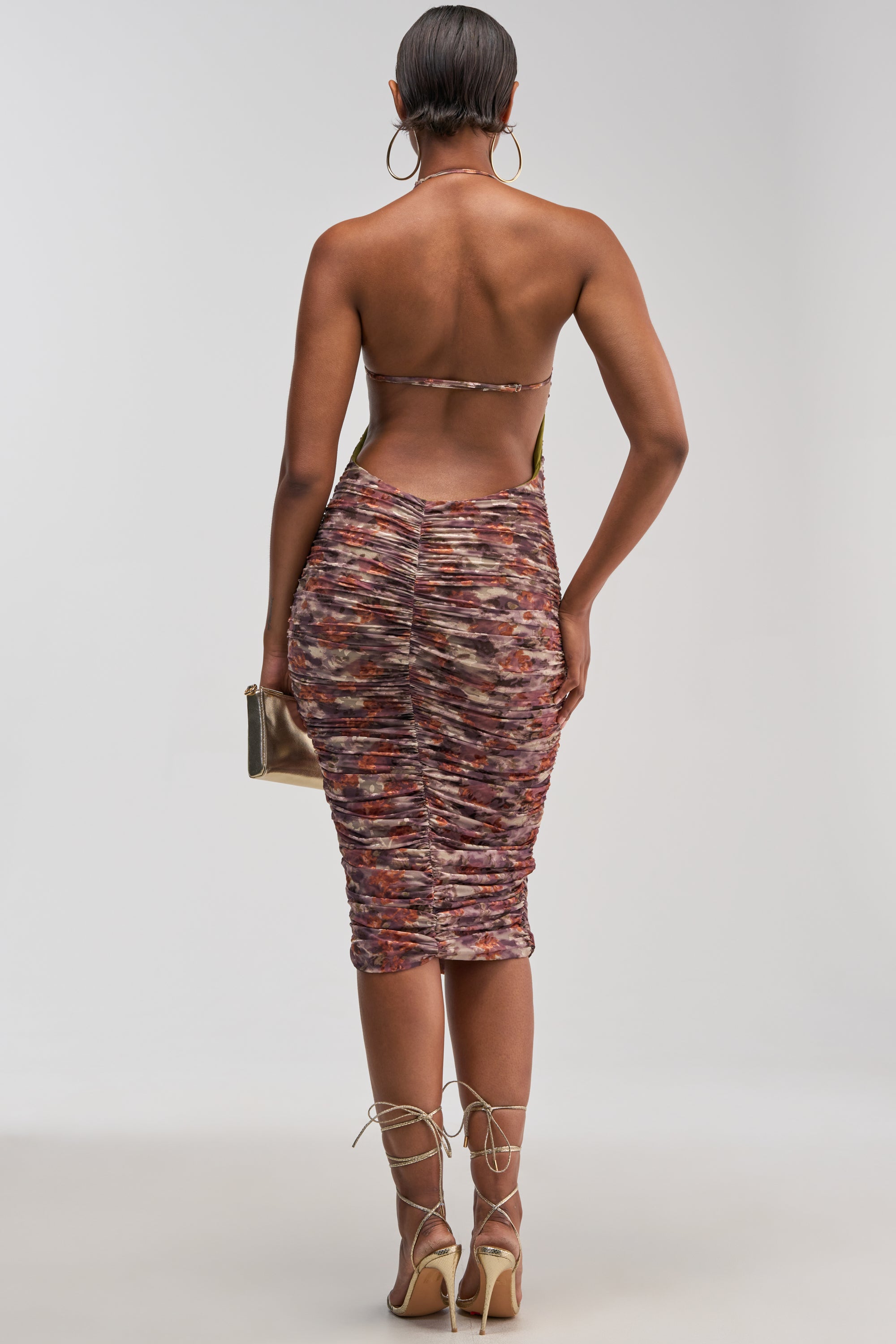 A woman stands facing away, wearing the SWEET AS CANDY FLORAL BURNOUT MIDI DRESS—a colorful, ruched midi dress featuring a backless halter neckline. She pairs it with strappy heels and hoop earrings, holding a small clutch against a plain background—a perfect choice for a wedding guest dress.