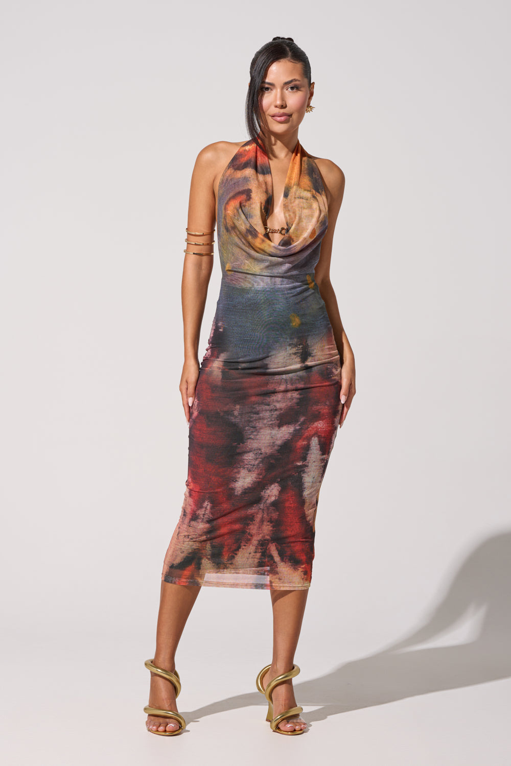Model wears the PICASSO DREAM GIRL PRINTED MIDI DRESS, a sleeveless midi dress featuring an abstract print, draped cowl neckline, and mid-length hem. She pairs the look with gold heels and a stack of gold bangles on one arm, her hair swept to one side against a simple backdrop—embodying evening elegance in this standout dress.