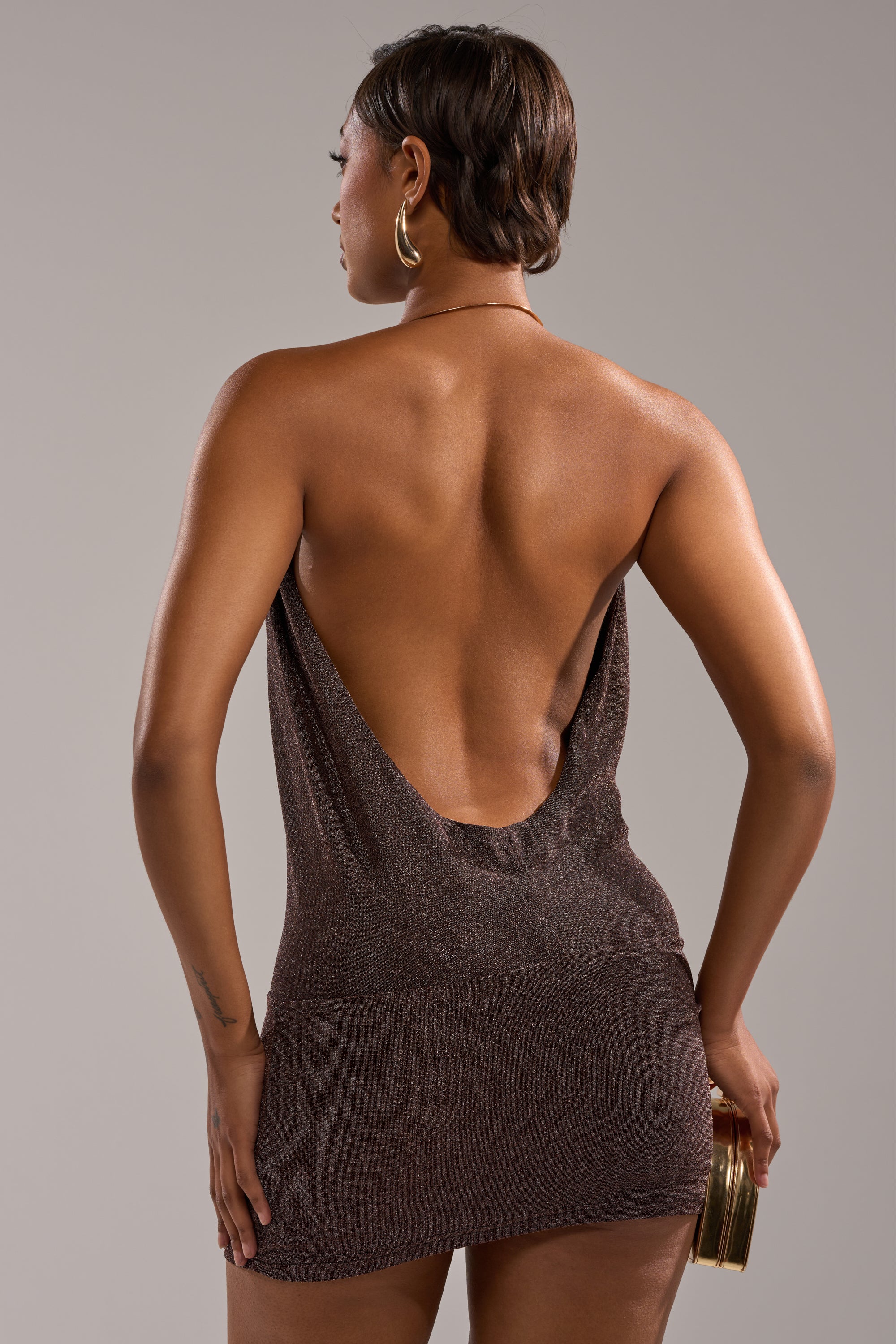 A person with short hair faces away from the camera against a grey background, wearing the VEGAS NIGHTS MINI DRESS—a shimmering, backless, body-skimming brown mini dress—and holding a gold clutch.