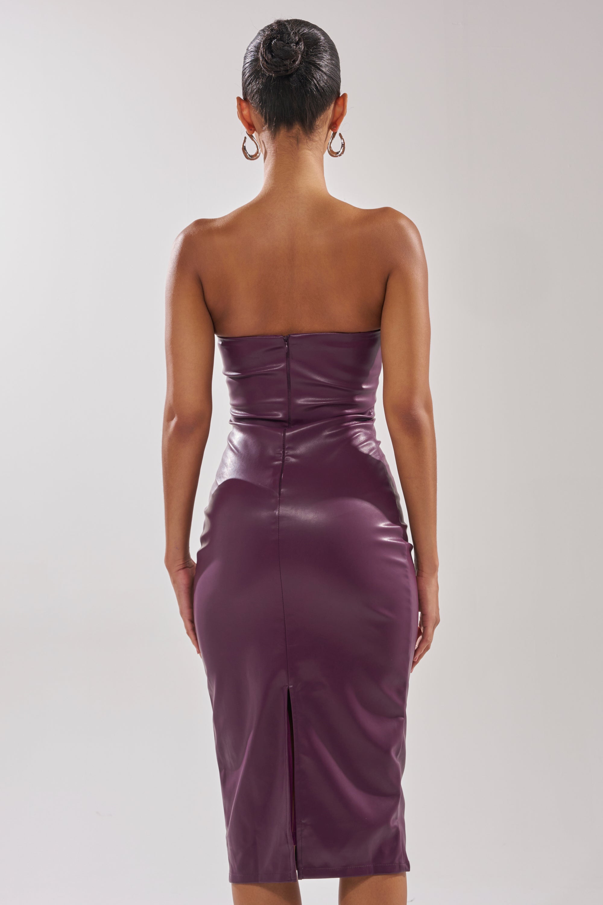 A woman faces away in the OH HOT DAMN FAUX LEATHER MIDI DRESS IN PURPLE, a strapless, curve-hugging style with a back slit. She wears her hair in a neat bun, hoop earrings, and stands against a plain light background. 6