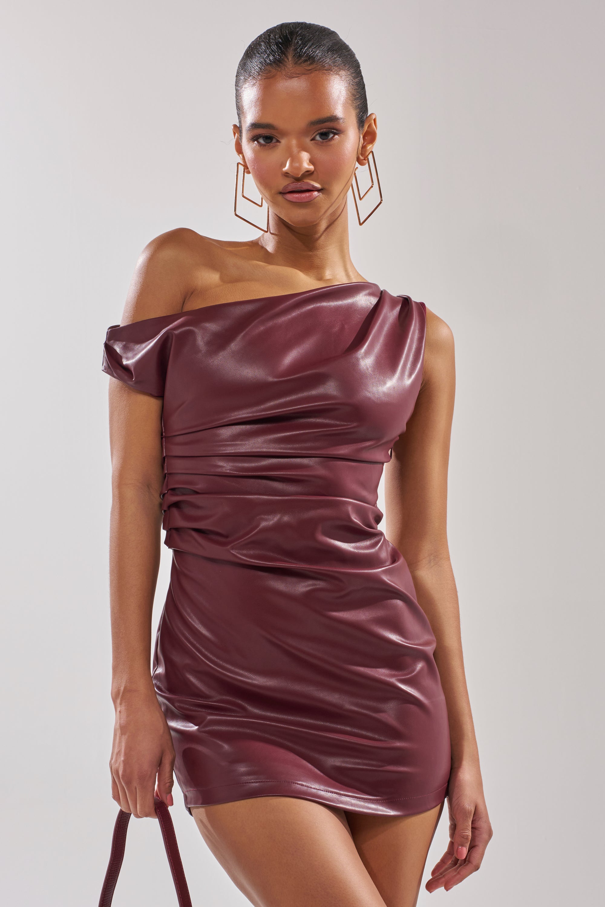 A woman wears the KYLIE PU MINI DRESS in shiny maroon with ruched details, holds a matching purse, has her hair pulled back, and accessorizes with large geometric earrings.