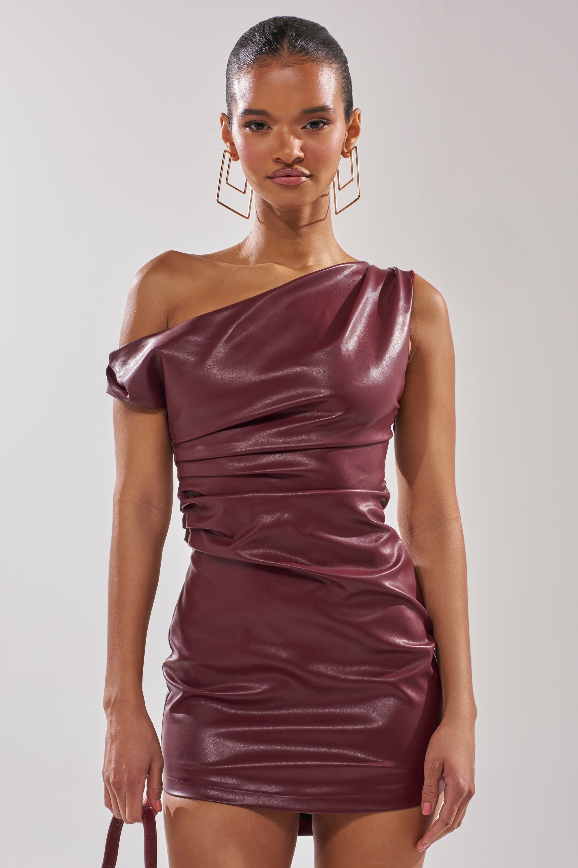 A woman stands against a plain background wearing the KYLIE PU MINI DRESS— a maroon, one-shoulder leather mini dress with ruched details. Her hair is sleek and pulled back, and she carries a matching bag.