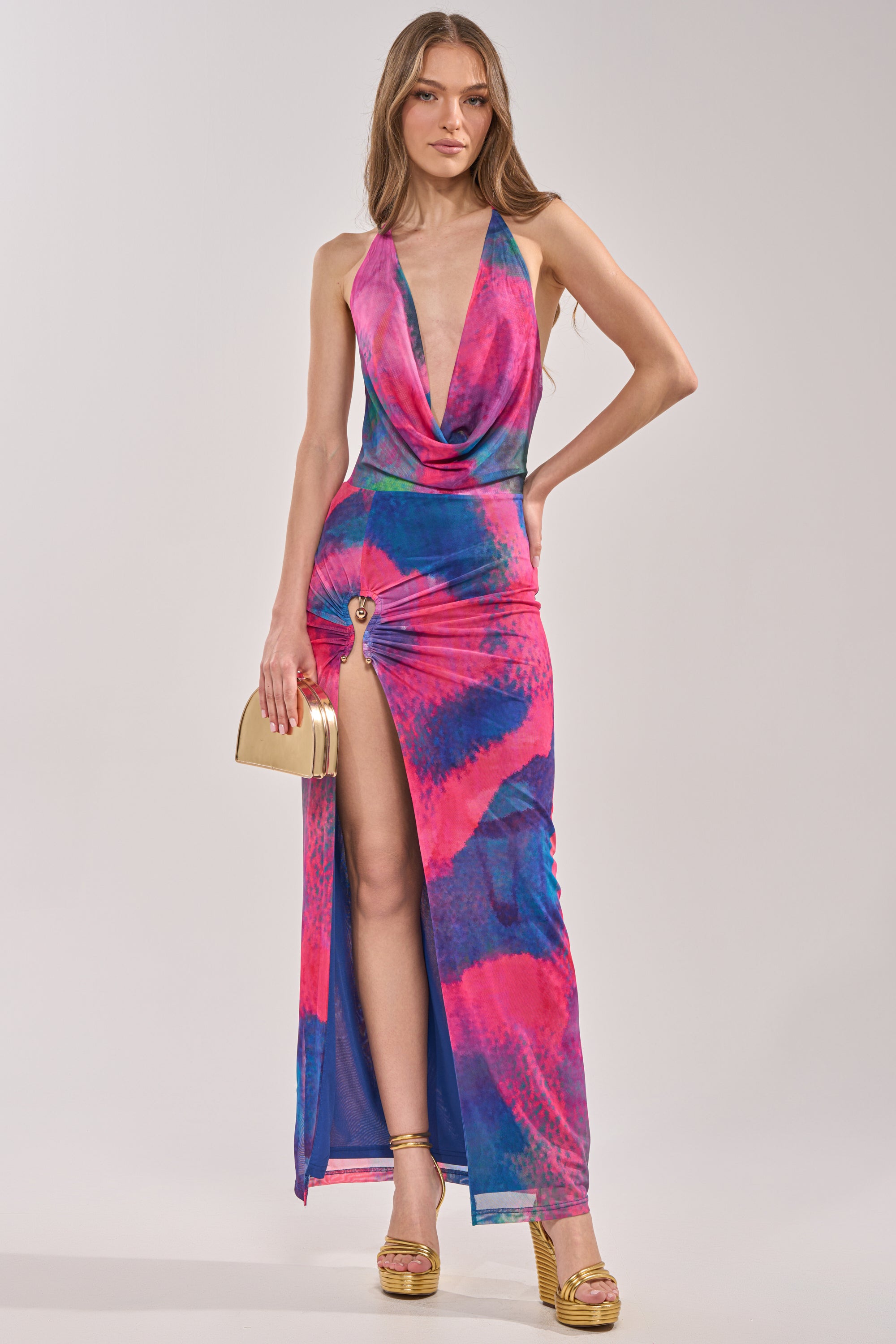 A woman in the ARTISTS MUSE MAXI DRESS, featuring a colorful tropical print, deep neckline, and high leg slit, pairs it with a gold clutch and gold platform heels against a plain light background. 3