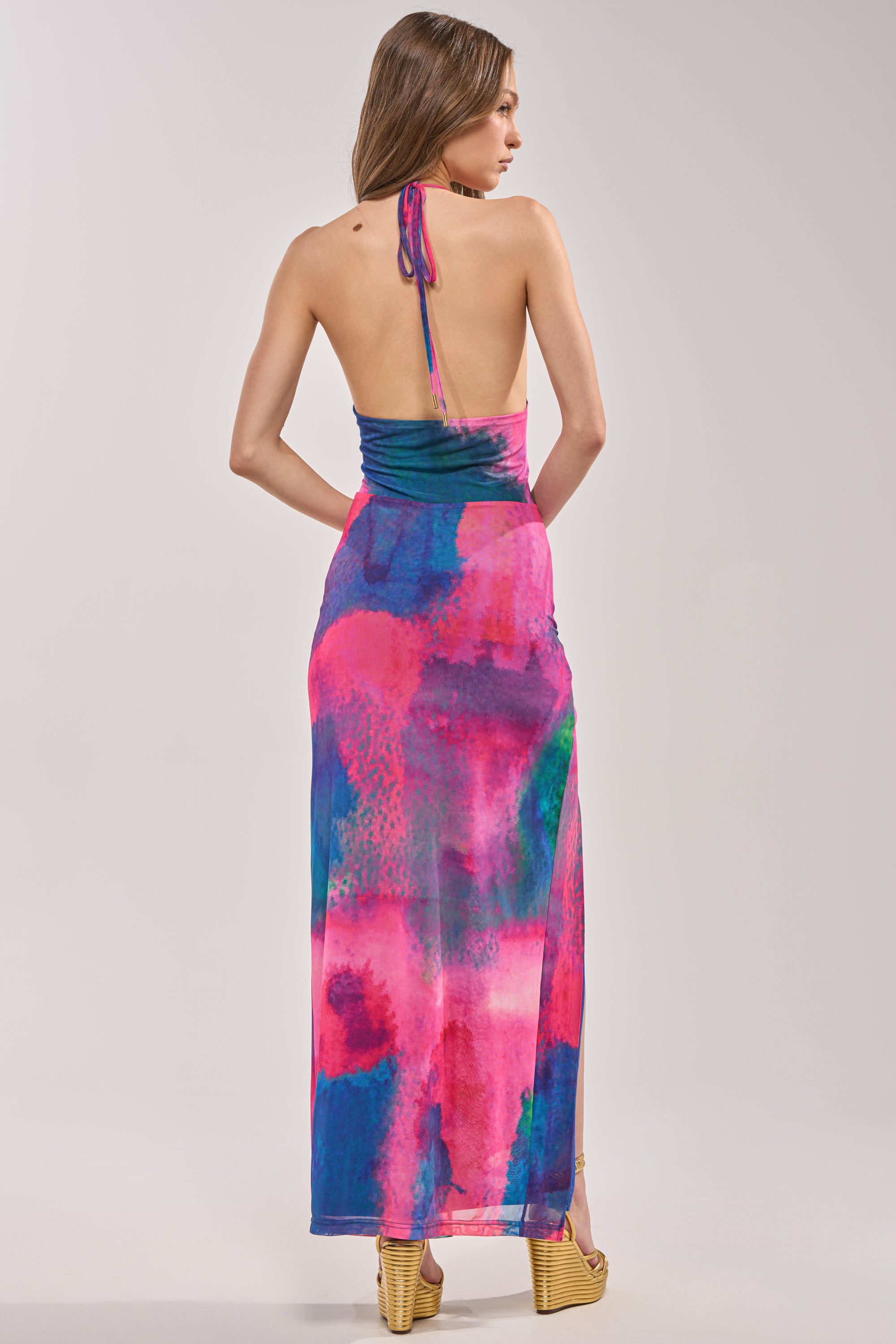 A woman with long hair and wedge sandals faces away, wearing the ARTISTS MUSE MAXI DRESS—a colorful tropical print halter maxi with pink, purple, blue, and green patterns—against a plain light gray background. 5