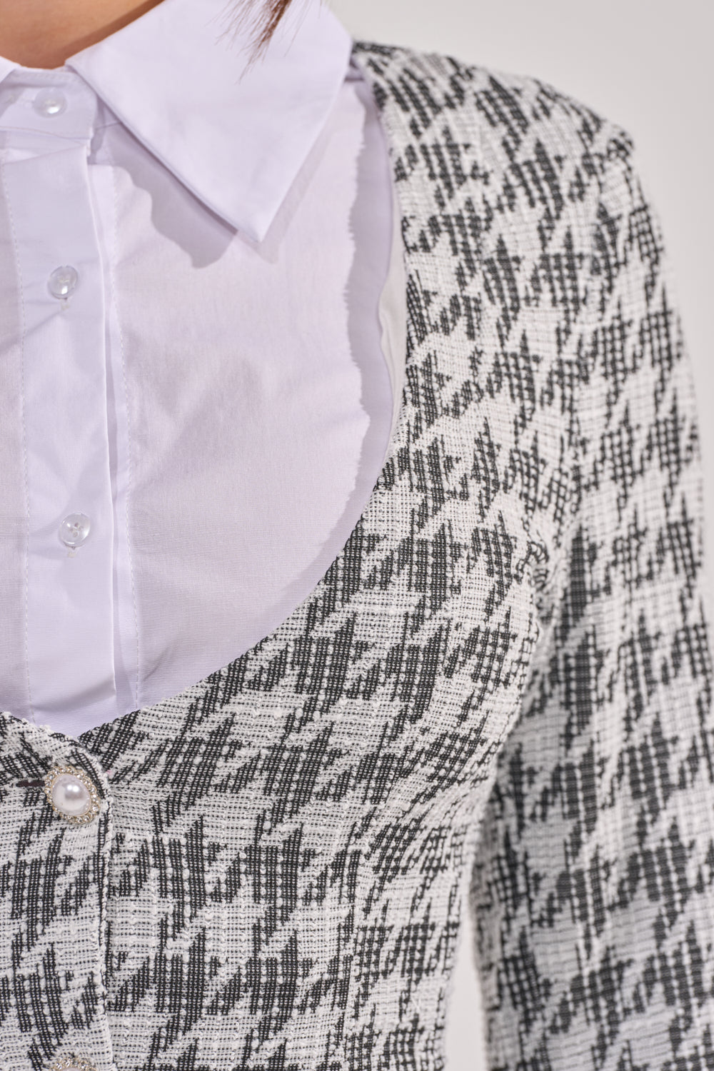 Close-up of a person wearing the TALIA TWEED LONG SLEEVE COLLARED DRESS under a black and white houndstooth blazer with a pearl button, highlighting the upper torso and details of the houndstooth blazer dress. 9