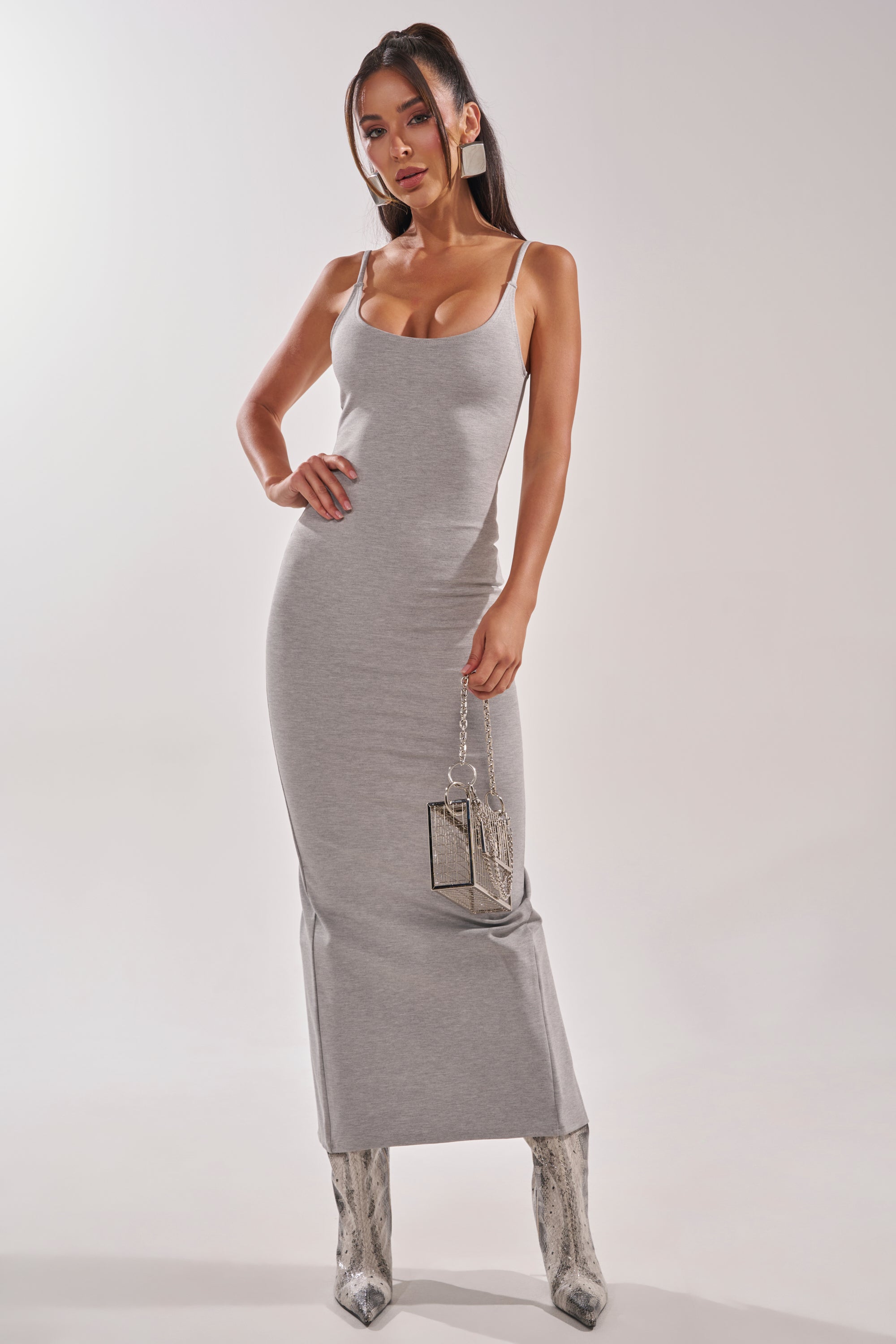A woman stands confidently in the fitted REYNA MAXI BODYCON DRESS IN GREY with thin straps, holding a small metallic purse and wearing pointed snakeskin-patterned boots against a plain light background. 1
