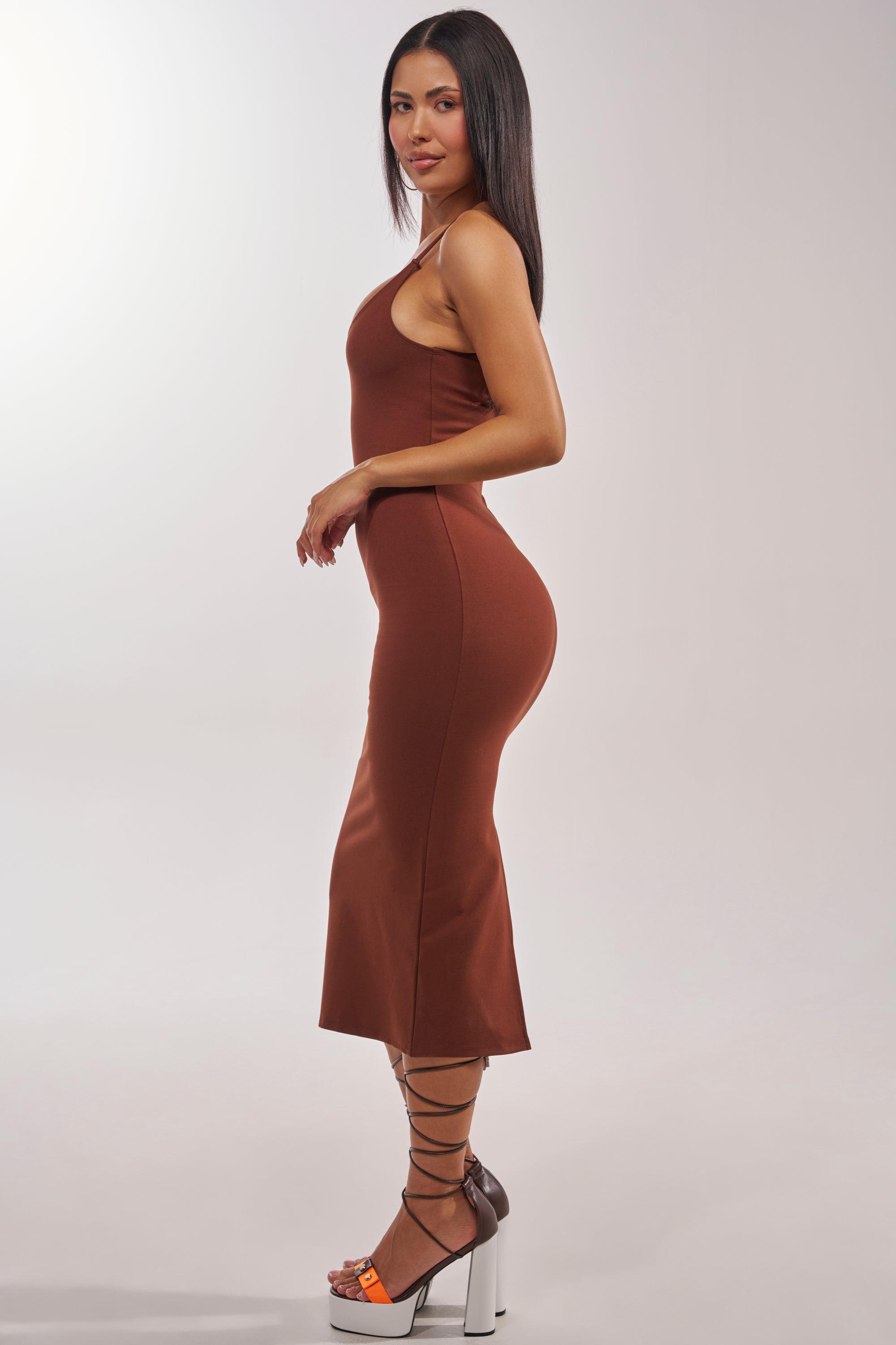 A woman stands in profile against a neutral background wearing the REYNA MIDI BODYCON DRESS IN BROWN with high-heeled sandals that feature black laces and orange accents. 2