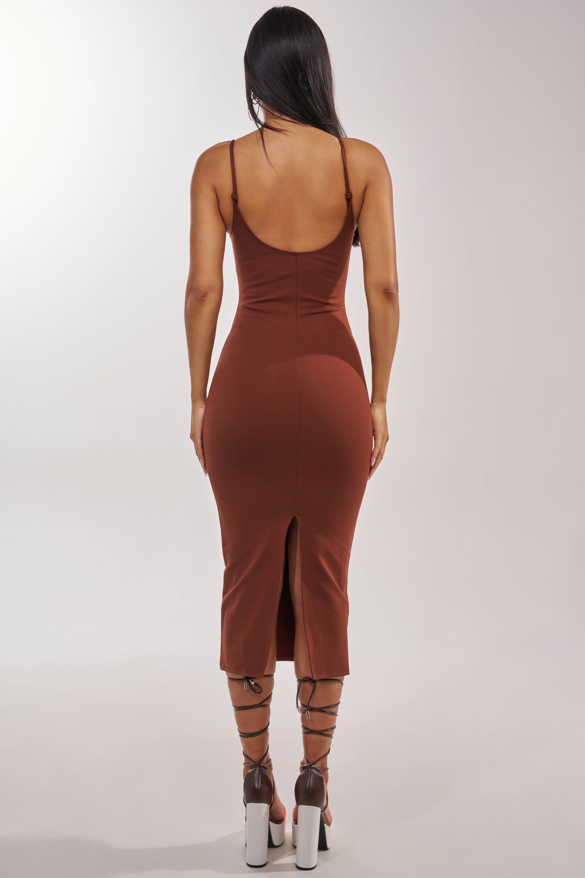 A woman with long dark hair models the REYNA MIDI BODYCON DRESS IN BROWN, featuring a back slit. She poses with her back to the camera against a plain background, highlighting this elegant evening dress. 3