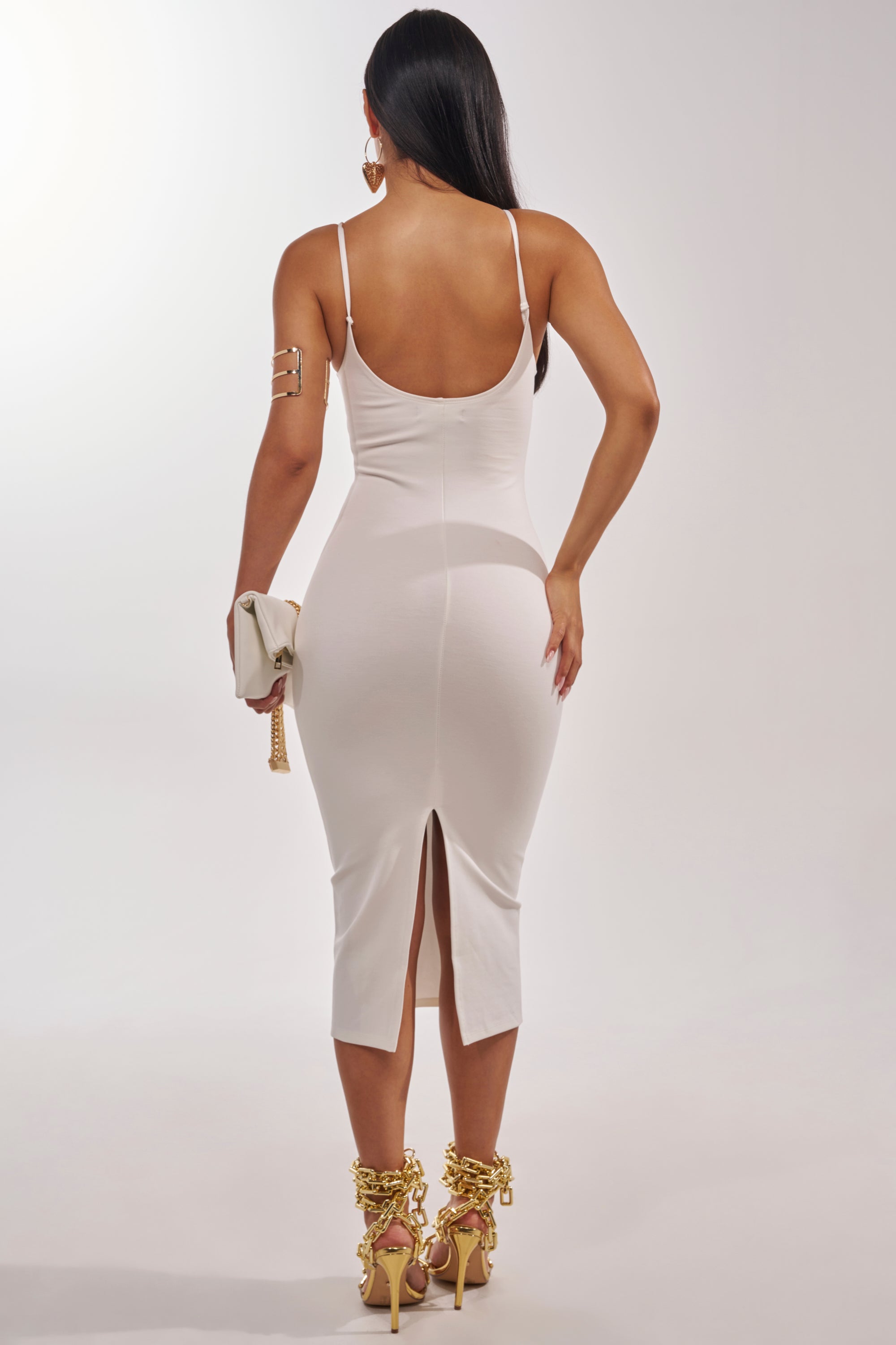 A woman faces away, wearing the REYNA MIDI BODYCON DRESS IN WHITE with spaghetti straps, paired with gold heels, gold jewelry, and holding a small white clutch against a plain light background. 4