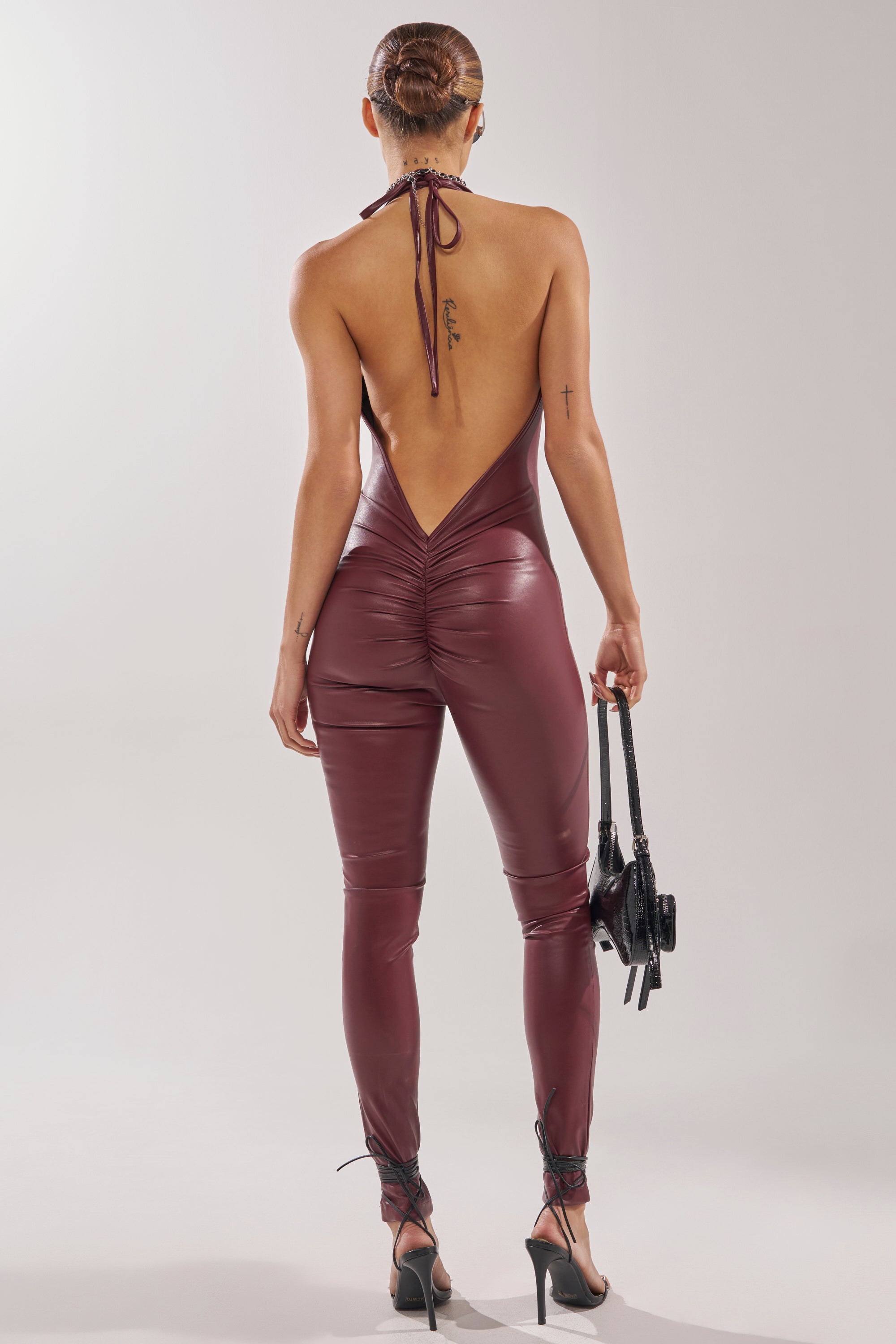 A person with a sleek bun wears the LATE TO THE PARTY LEATHER JUMPSUIT IN BURGUNDY—club-ready, backless, halter style—paired with high heels and a small black handbag, standing against a plain light background. 5
