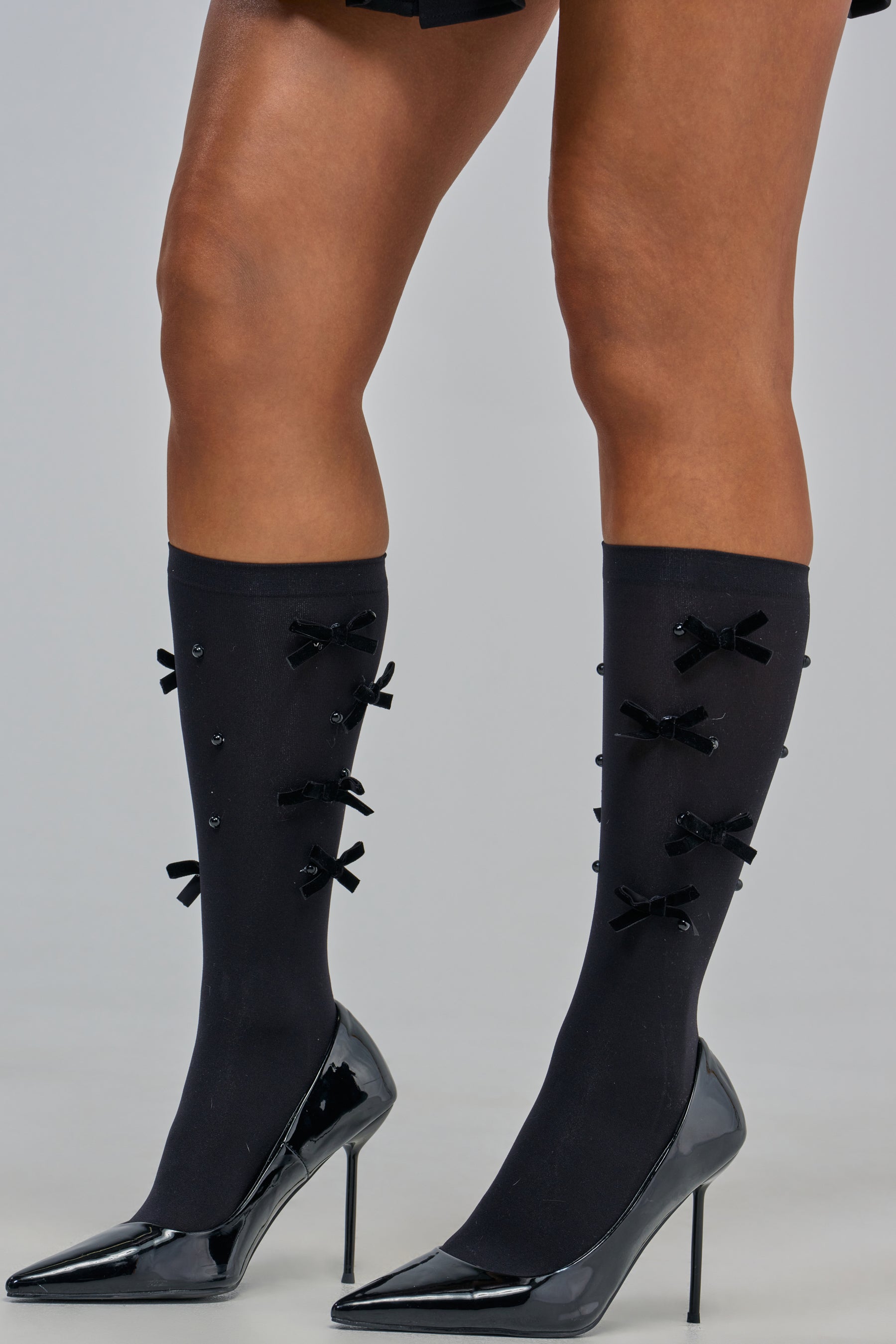 Model wearing DOLLCORE RIBBON CREW SOCKS—black socks adorned with small bows—paired with shiny black high-heeled shoes, standing on a light gray background for a charming coquette-inspired look.