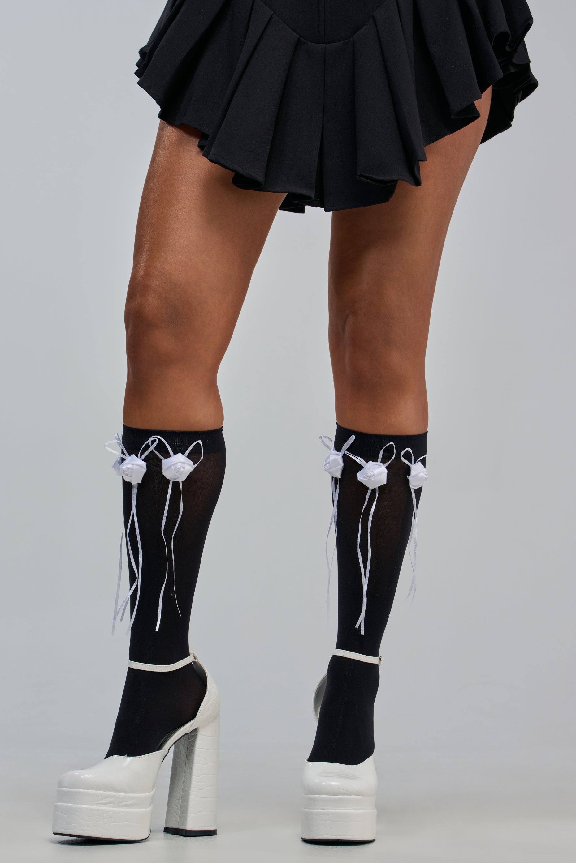 A person wearing BLACK ROSETTE SOCKS styled with a black pleated skirt and white high-heeled platform shoes, standing against a plain background.