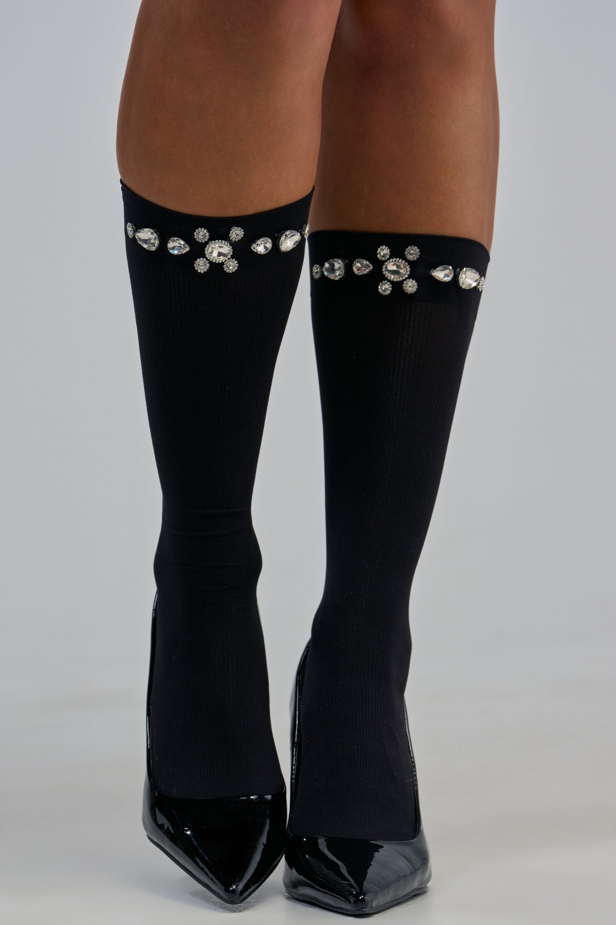A person wearing the JEWELED EDGE SOCKS IN CLEAR CRYSTAL, featuring a row of rhinestone details near the top, paired with shiny black pointed-toe high heels.
