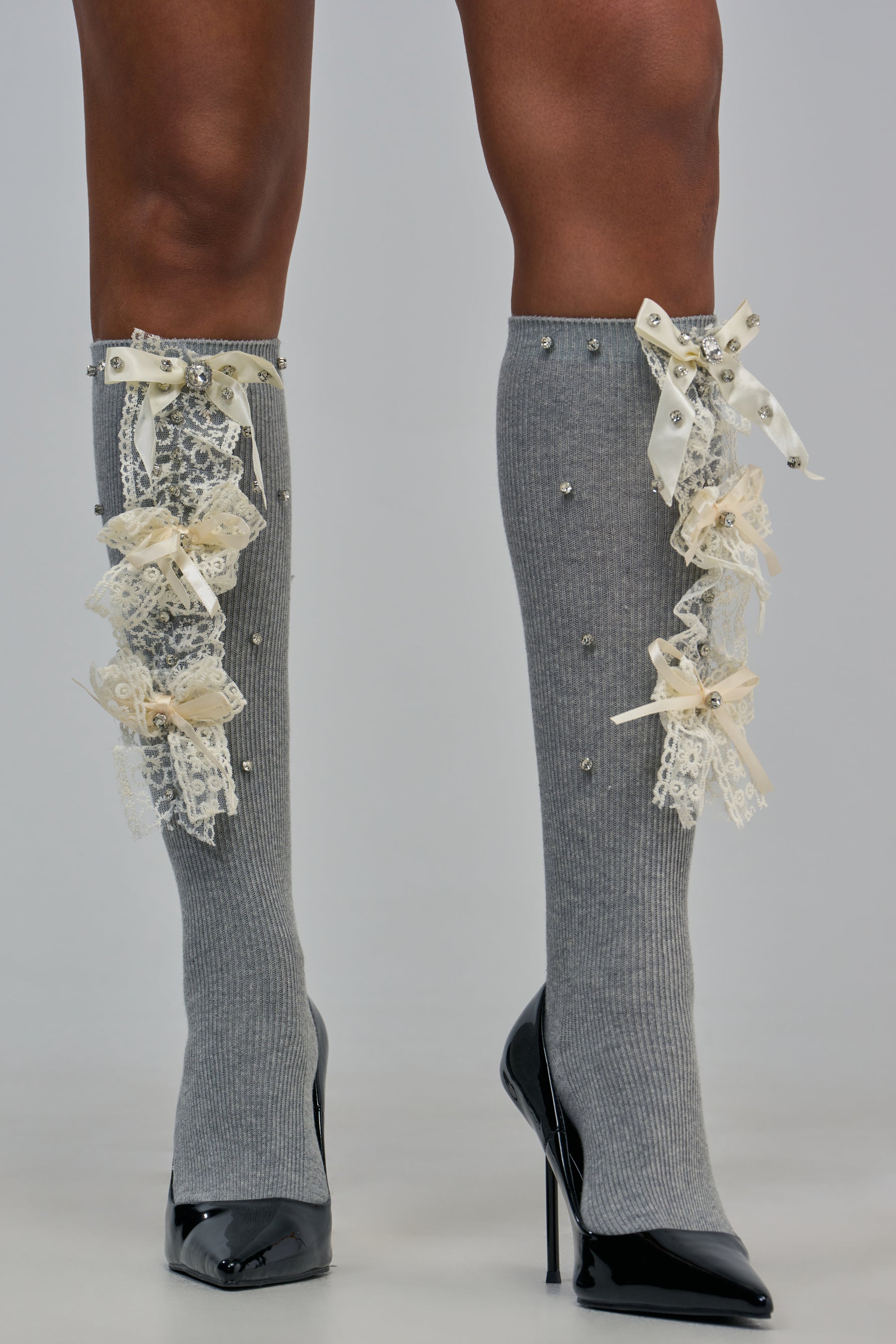 VINTAGE LACE BOW EMBELLISHED SOCKS in grey, featuring a ribbed knee-high design with cream lace bows and scattered rhinestones, styled with black high-heeled pumps for a vintage look.