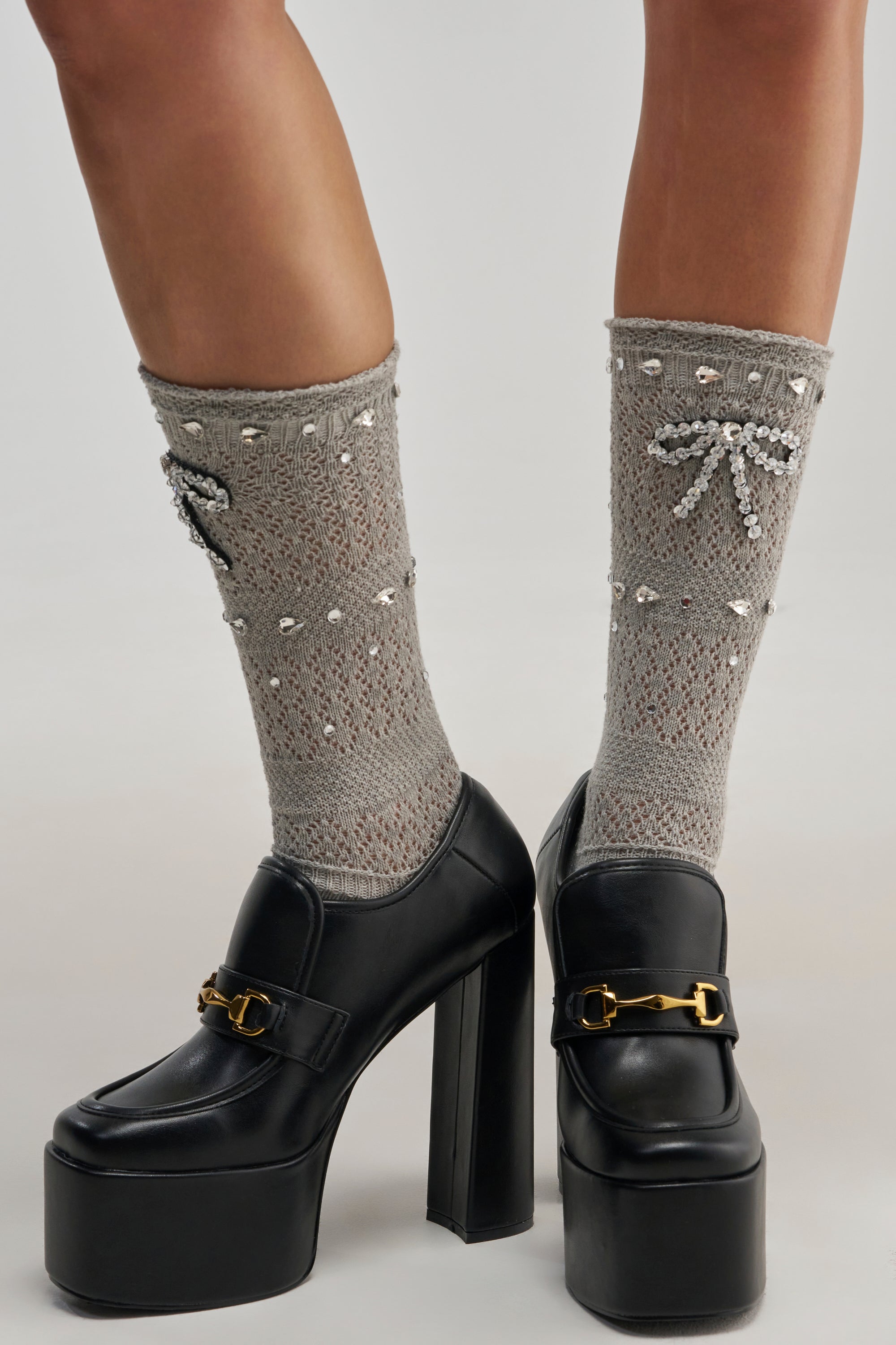 Wearing the ICED OUT BOW DETAIL SOCKS—light gray lace socks adorned with bows and small pearl accents—paired with black platform heels featuring gold buckle details, creates a charming school girl chic look.