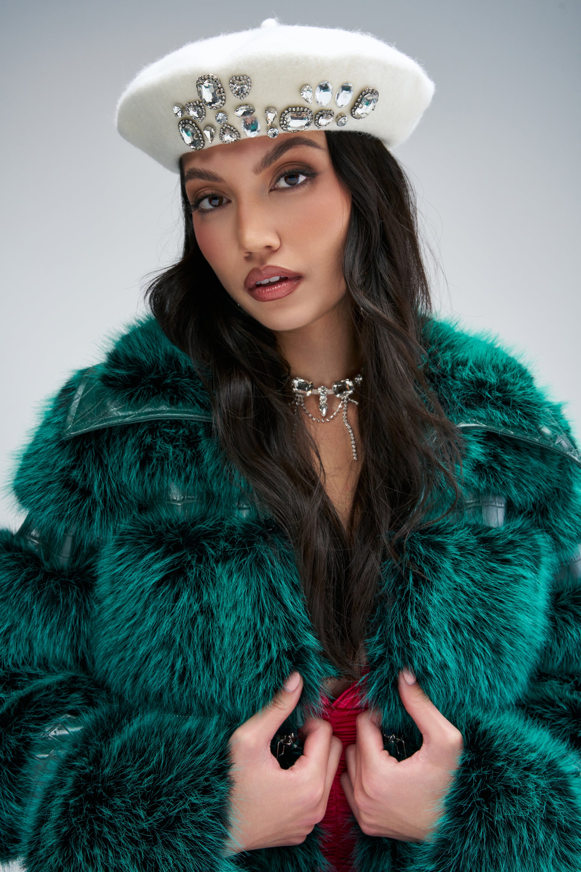 A woman wears a white CREME DE LA CREME BERET decorated with large crystals and a vibrant green faux fur coat.