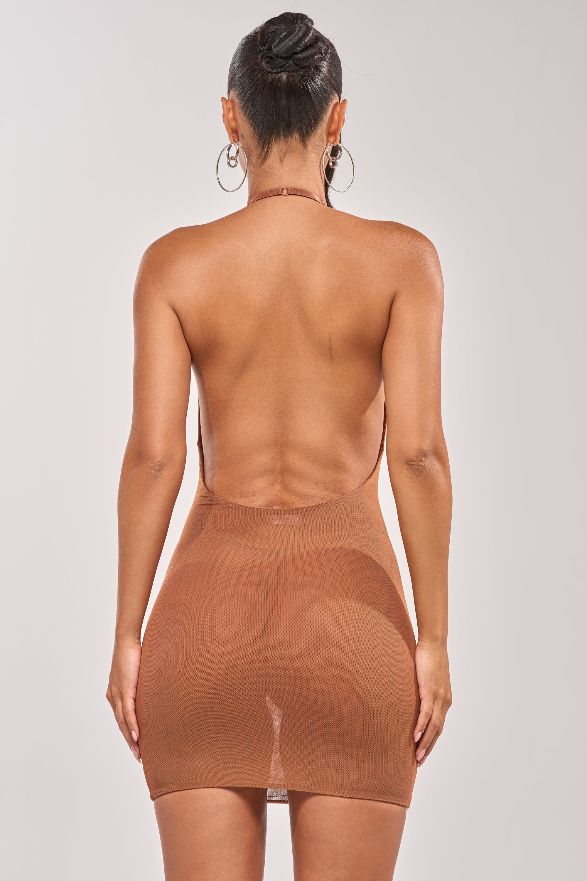A woman with her hair in a bun and large hoop earrings models the BARELY THERE MESH MINI DRESS IN BROWN, a backless halter-neck mini, standing with her back to the camera against a plain background—showcasing this layering essential. 4