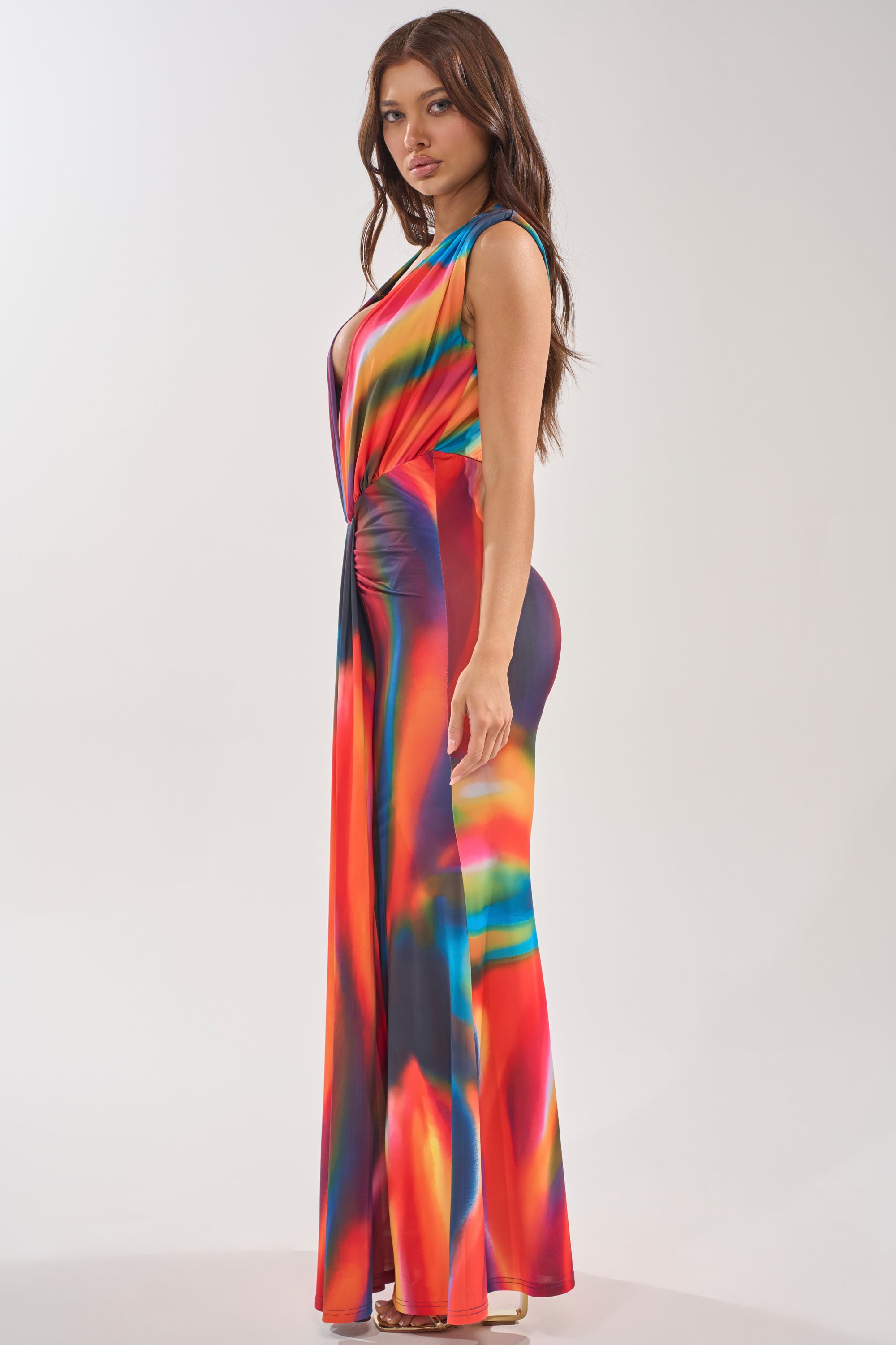 Woman standing sideways in the "Color Me Impressed Maxi Dress," featuring a vivid abstract watercolor print and wide flared silhouette, paired with open-toe heels against a plain light background. 3