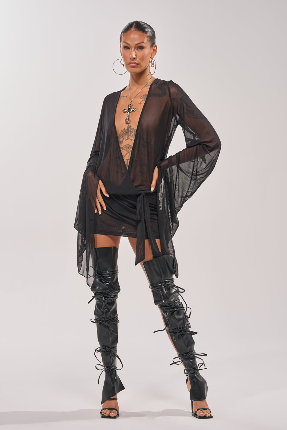 A person poses confidently in the SHEER AFFECTION MESH MINI DRESS with thigh-high lace-up boots, tattoos, and hoop earrings, creating a bold, stylish look against a plain light background. 1