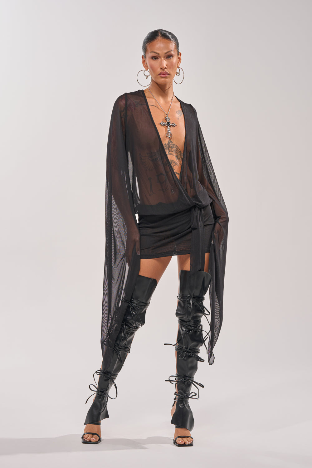 A woman stands confidently in the SHEER AFFECTION MESH MINI DRESS with flowing sleeves, tattoos, large hoop earrings, a cross necklace, and lace-up thigh-high boots against a plain light background—ideal for evening wear. 4