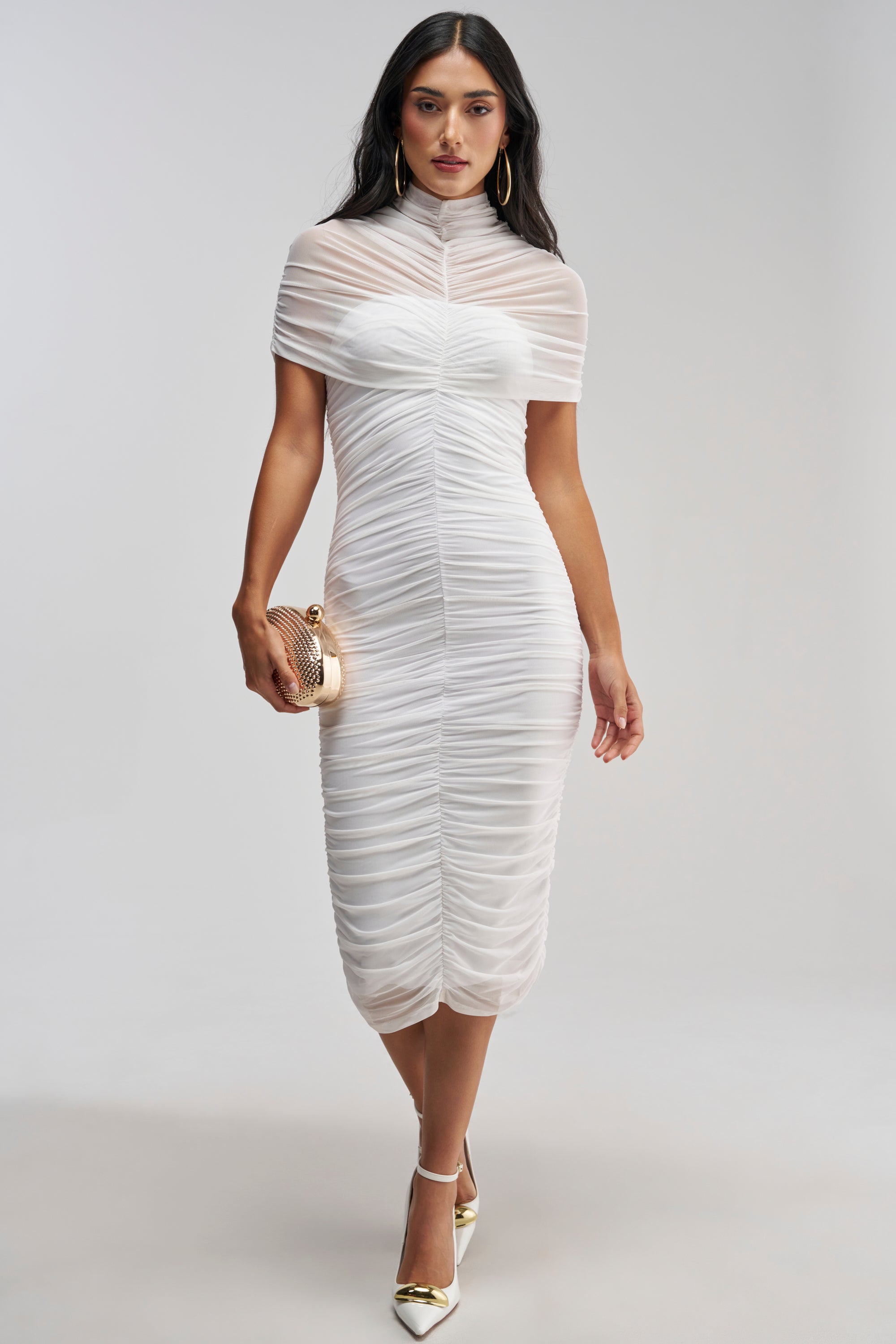 Dressed in the FIRST CLASS FANTASY MESH MIDI DRESS, this woman pairs her white, ruched, short-sleeve midi with a gold clutch, gold hoop earrings, and white heels featuring gold tips—an ideal ensemble for bridal events or any formal occasion.