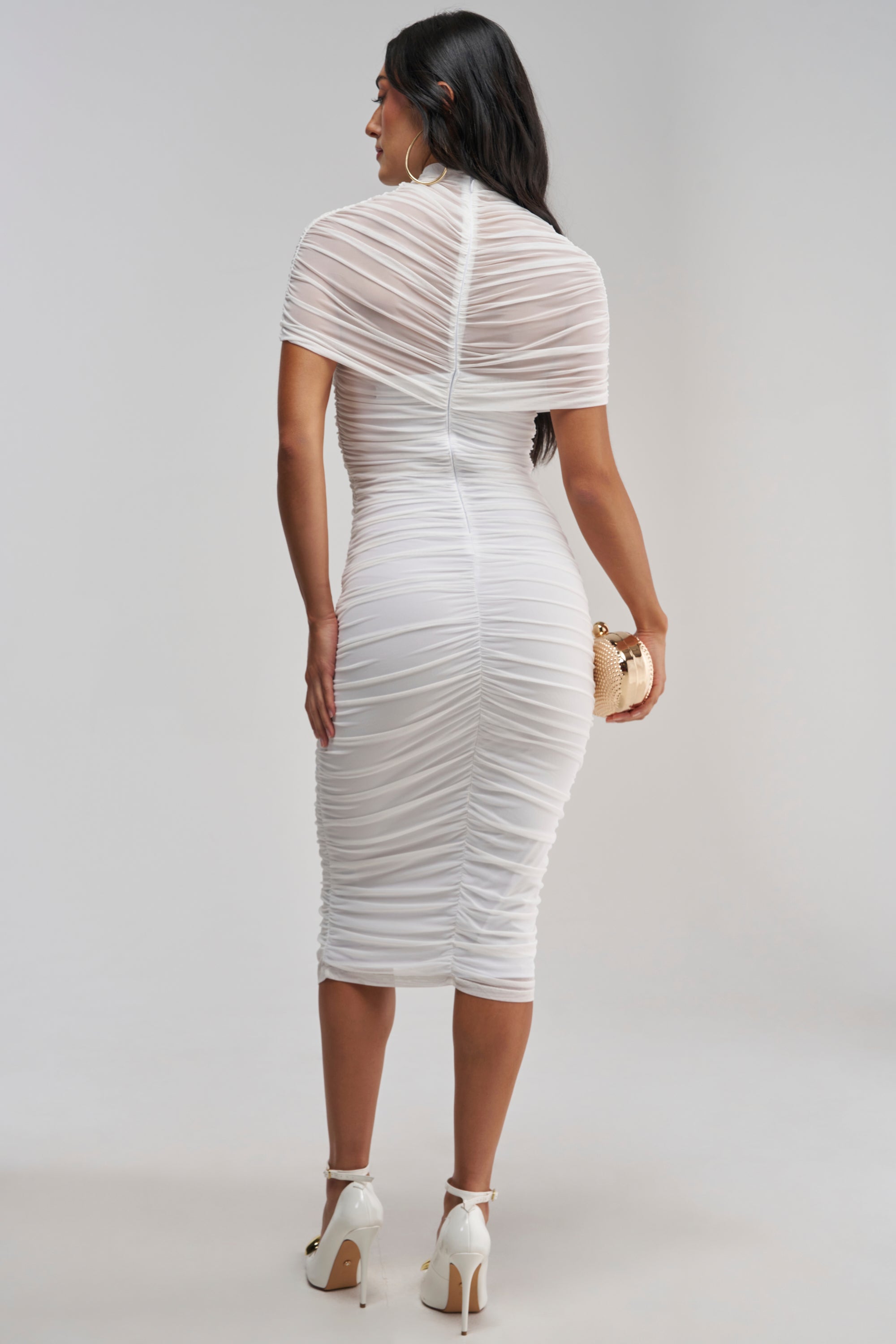 Facing away, a woman showcases the FIRST CLASS FANTASY MESH MIDI DRESS—a fitted white ruched midi dress ideal for bridal events or any formal occasion. She carries a small round purse, her long dark hair flowing down her back, and completes the look with white high heels against a simple light background.