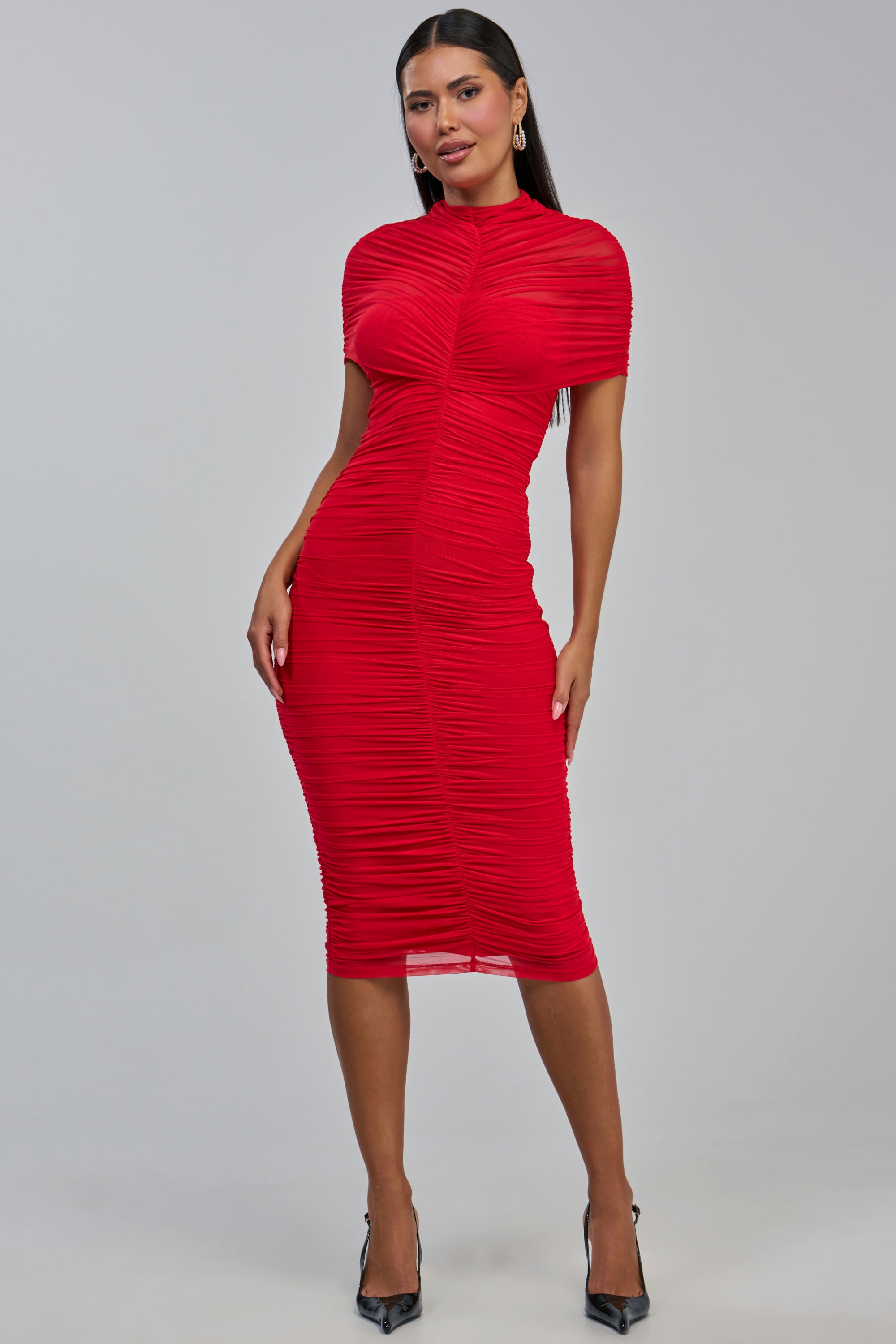 A woman models the FIRST CLASS FANTASY MESH MIDI DRESS IN RED, a fitted, ruched bodycon cocktail dress with short sleeves, paired with black heels against a plain light-colored background.