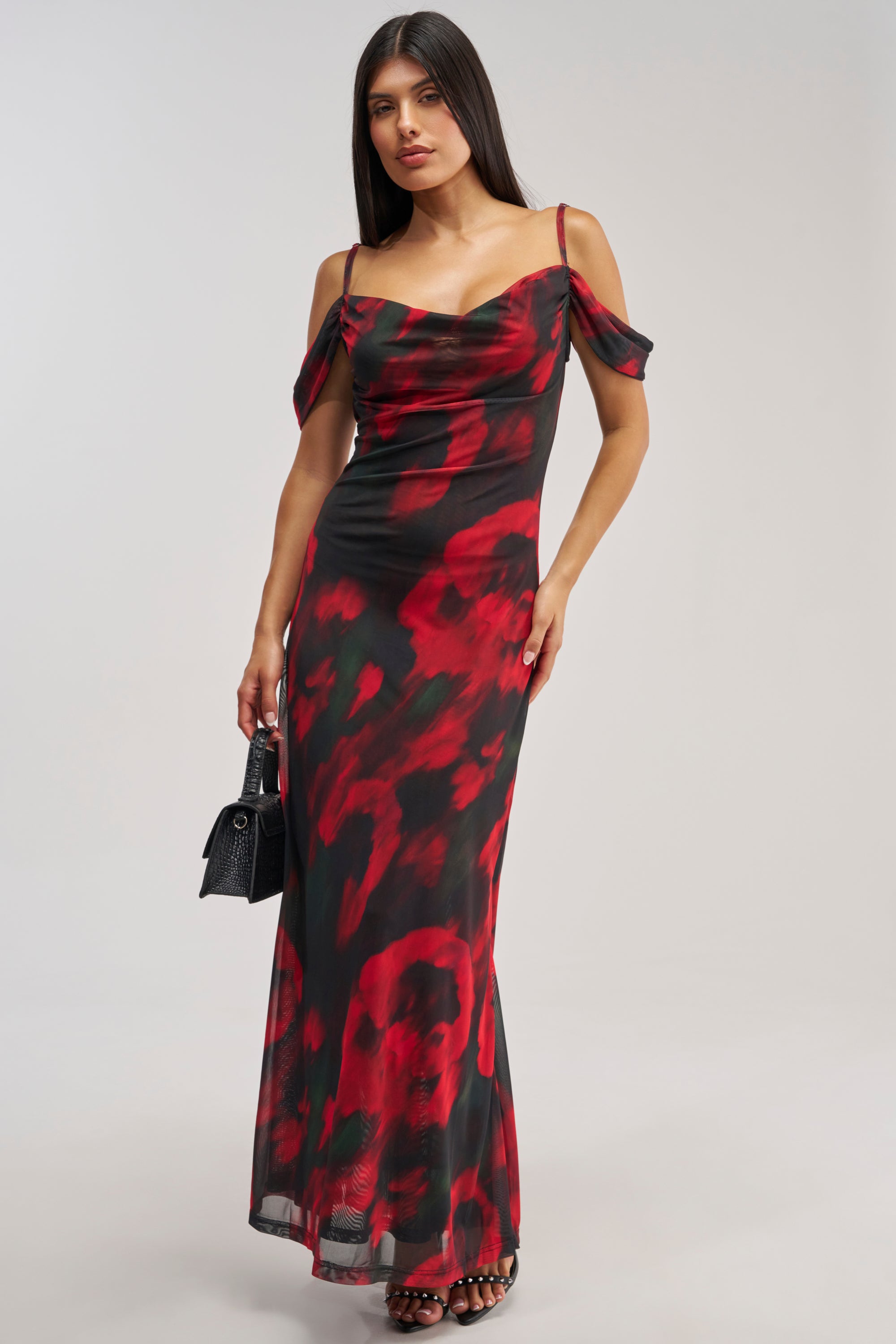 Dressed in the MIDNIGHT LOVER ABSTRACT MESH MAXI DRESS, a black off-the-shoulder mesh maxi adorned with red floral patterns, the woman holds a small black handbag and stands against a simple light background.