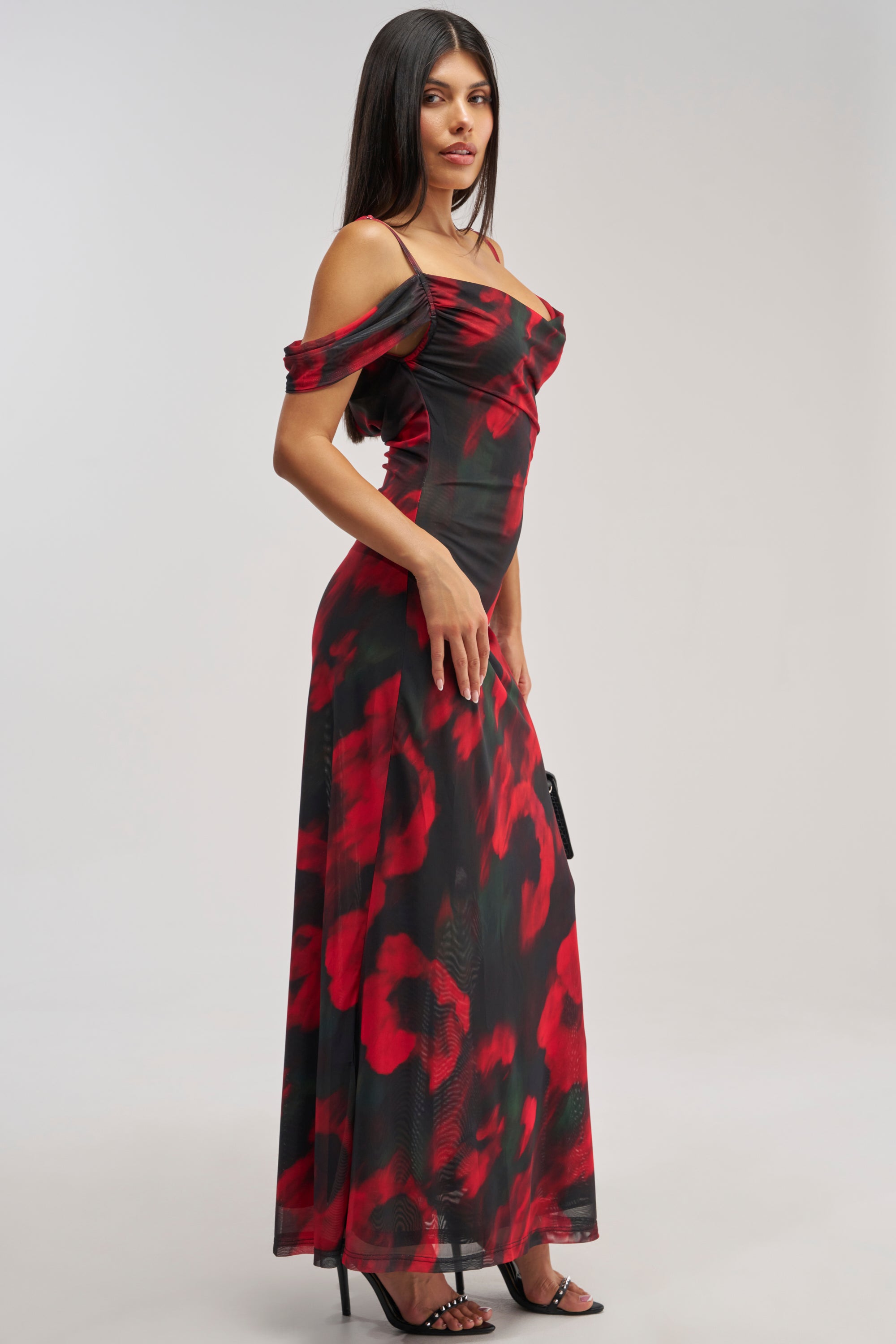 Wearing the MIDNIGHT LOVER ABSTRACT MESH MAXI DRESS—an off-the-shoulder black mesh maxi dress featuring bold red floral patterns—a woman stands in profile against a plain light background, paired with black high heels and sleek straight dark hair, making this an elegant option for any formal occasion or as a wedding guest dress.