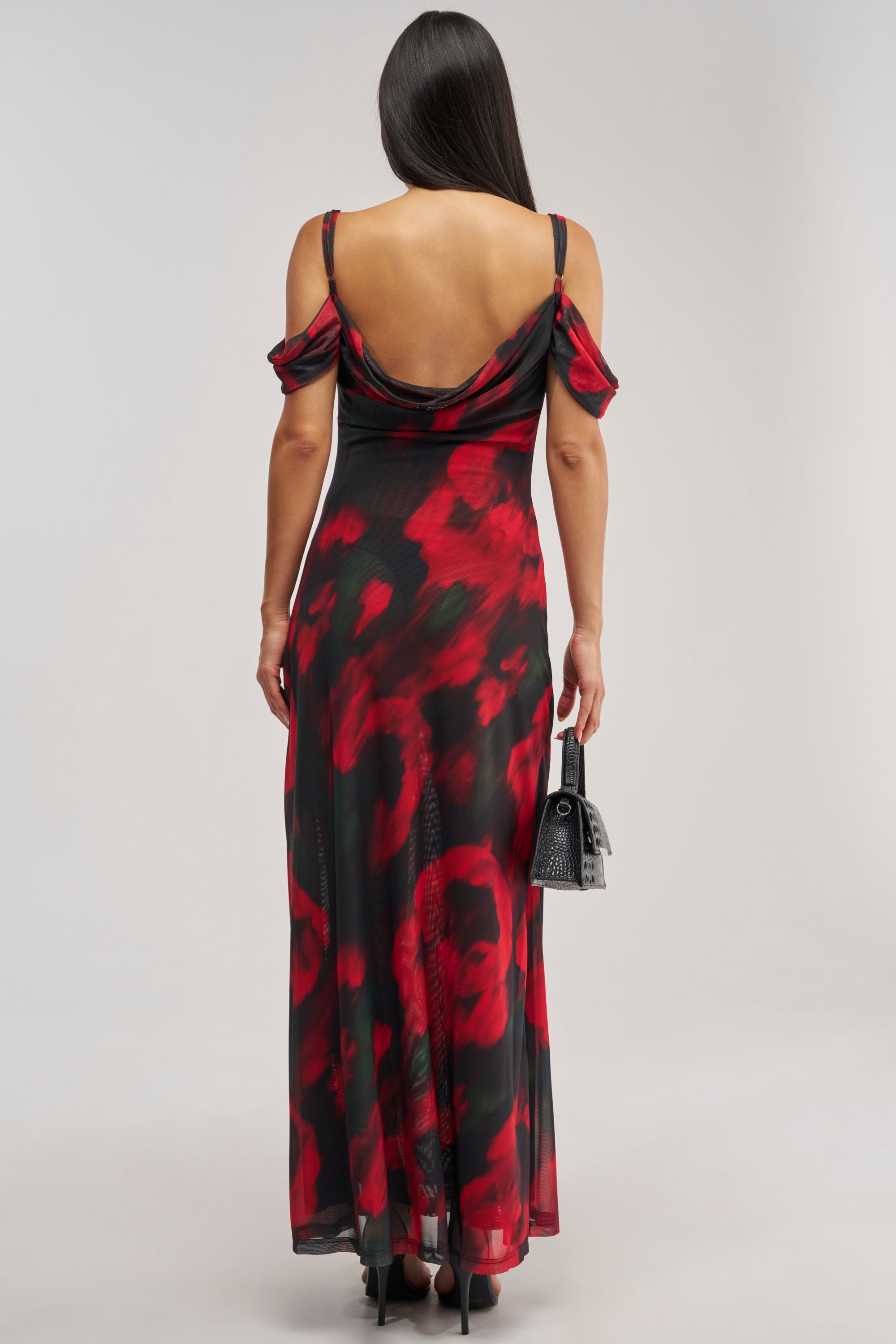 A woman with long, dark hair is seen from behind wearing the MIDNIGHT LOVER ABSTRACT MESH MAXI DRESS—a striking backless, red and black floral mesh off-the-shoulder maxi dress with thin straps. She holds a small black handbag.