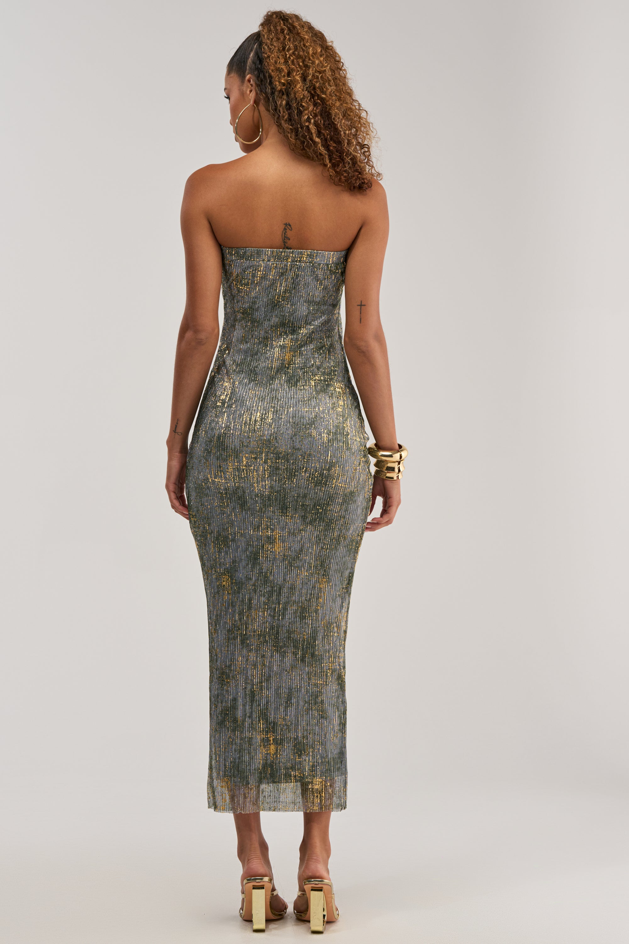 A woman with curly hair stands with her back to the camera, wearing the CHAMPAGNE GLOW STRAPLESS MAXI DRESS and gold heels. She has tattoos on her arms and back and accessorizes with gold bracelets.